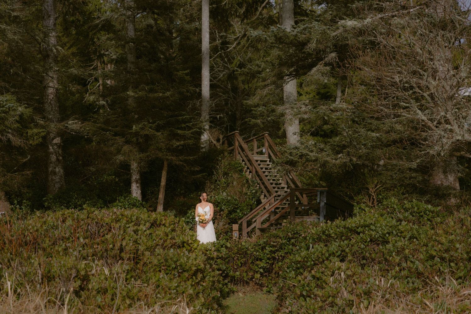 White wedding dress against lush forest backdrop with rustic wooden stairs.