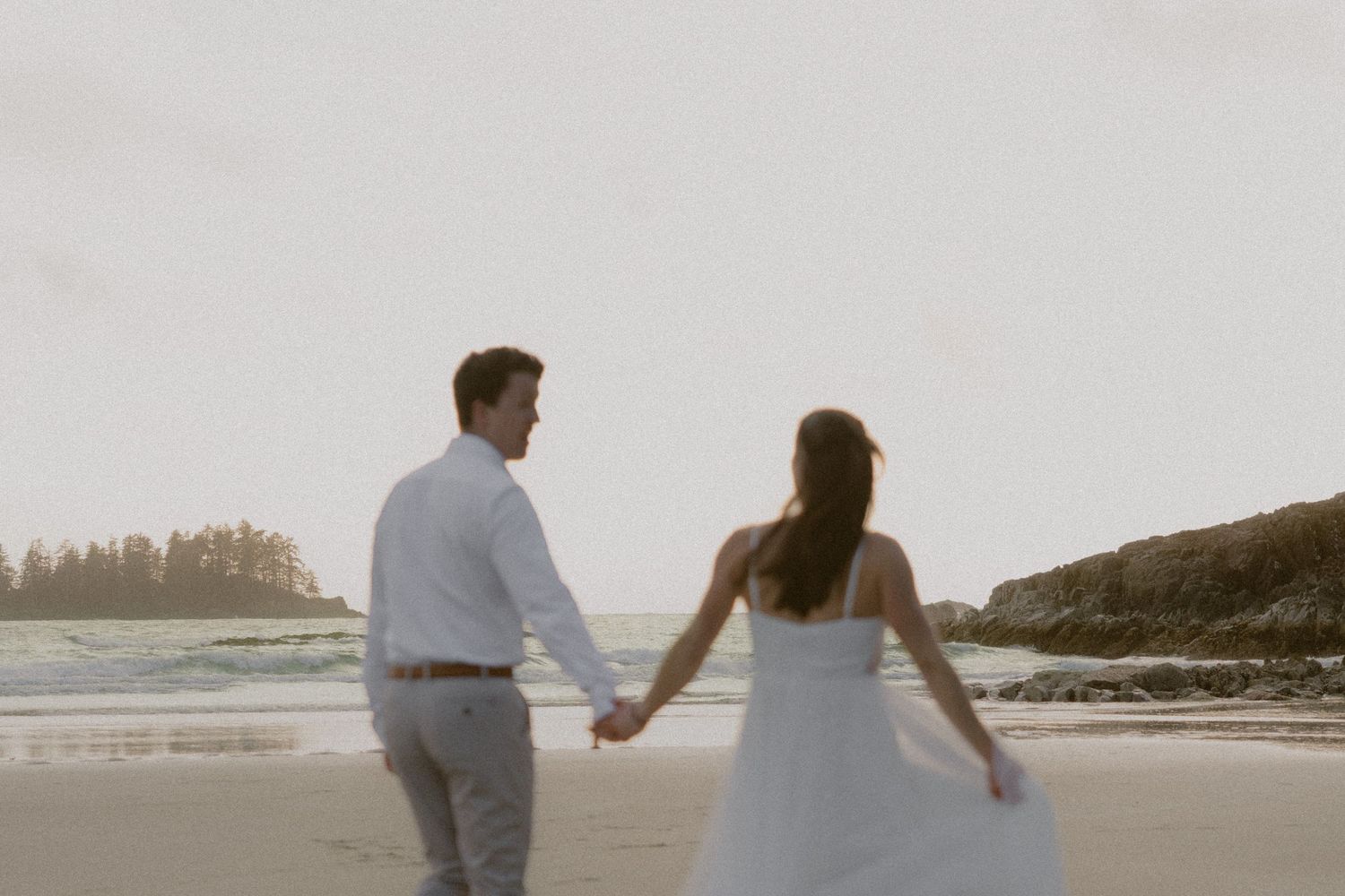 Couple strolling hand in hand on a scenic beach with cliffs and trees in the background.