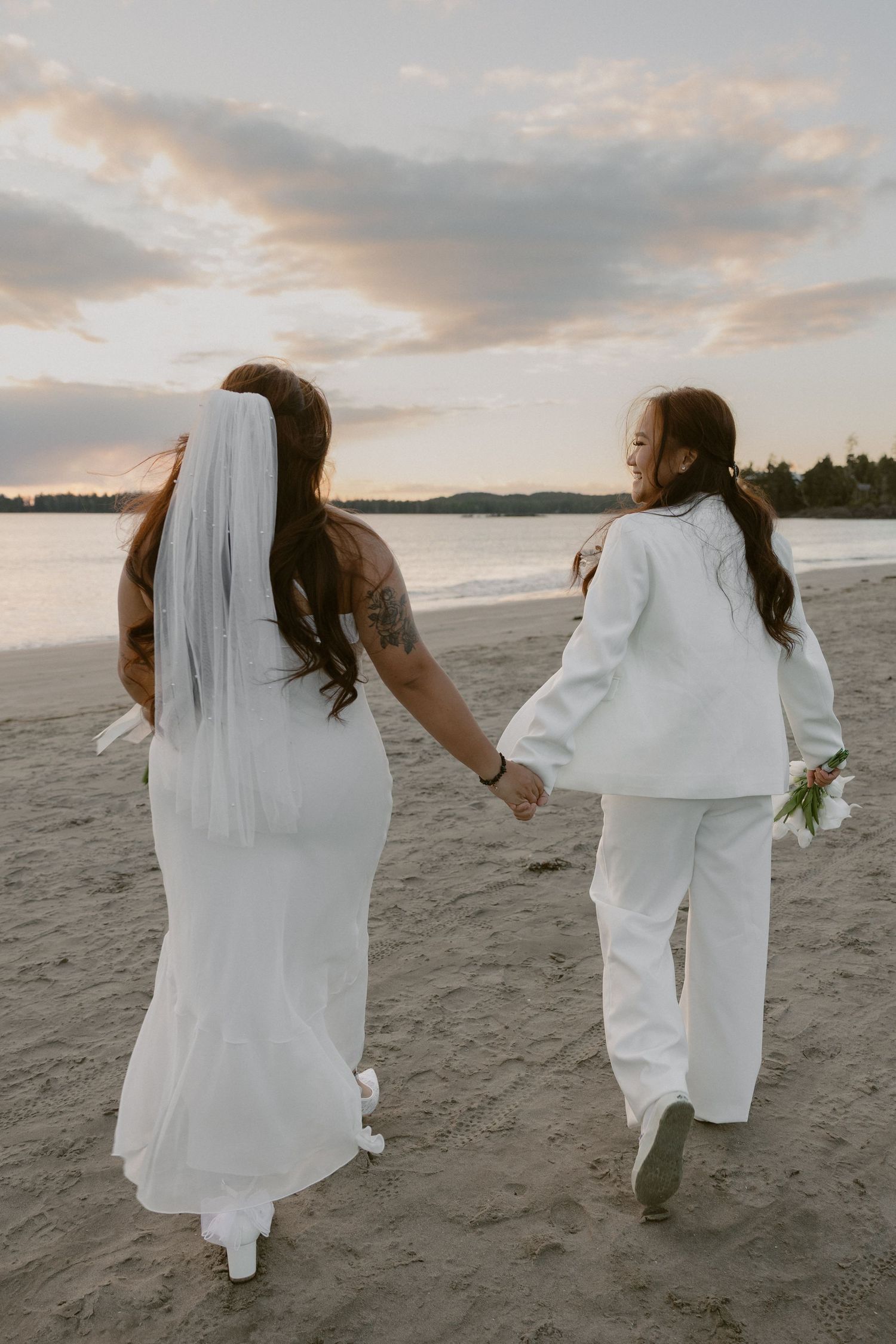 Two people in white dresses walking together on a beach during sunset.