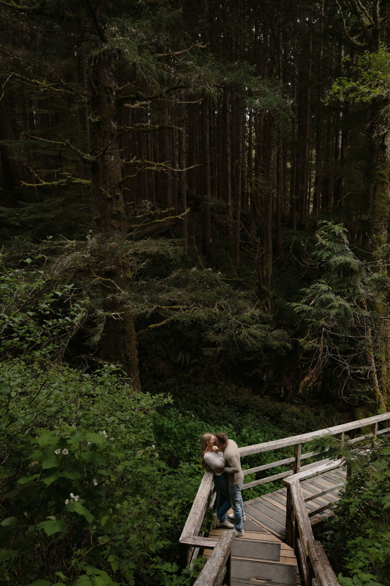 Couple embracing on a wooden bridge surrounded by lush green forest trees in a romantic outdoor setting.