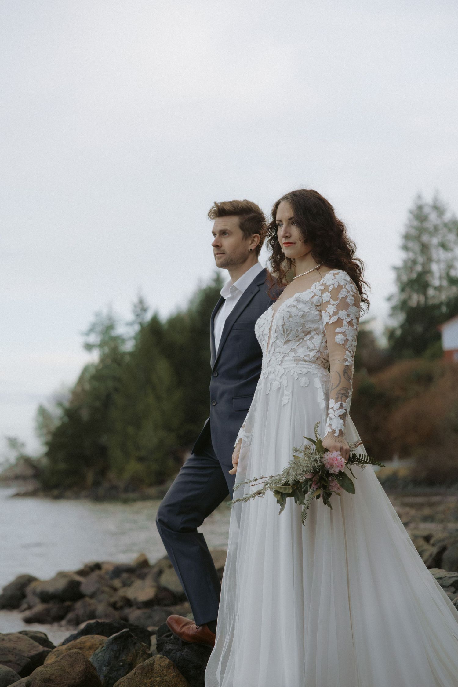 A couple shares an intimate moment on a rocky shoreline with mountains and water in the background.
