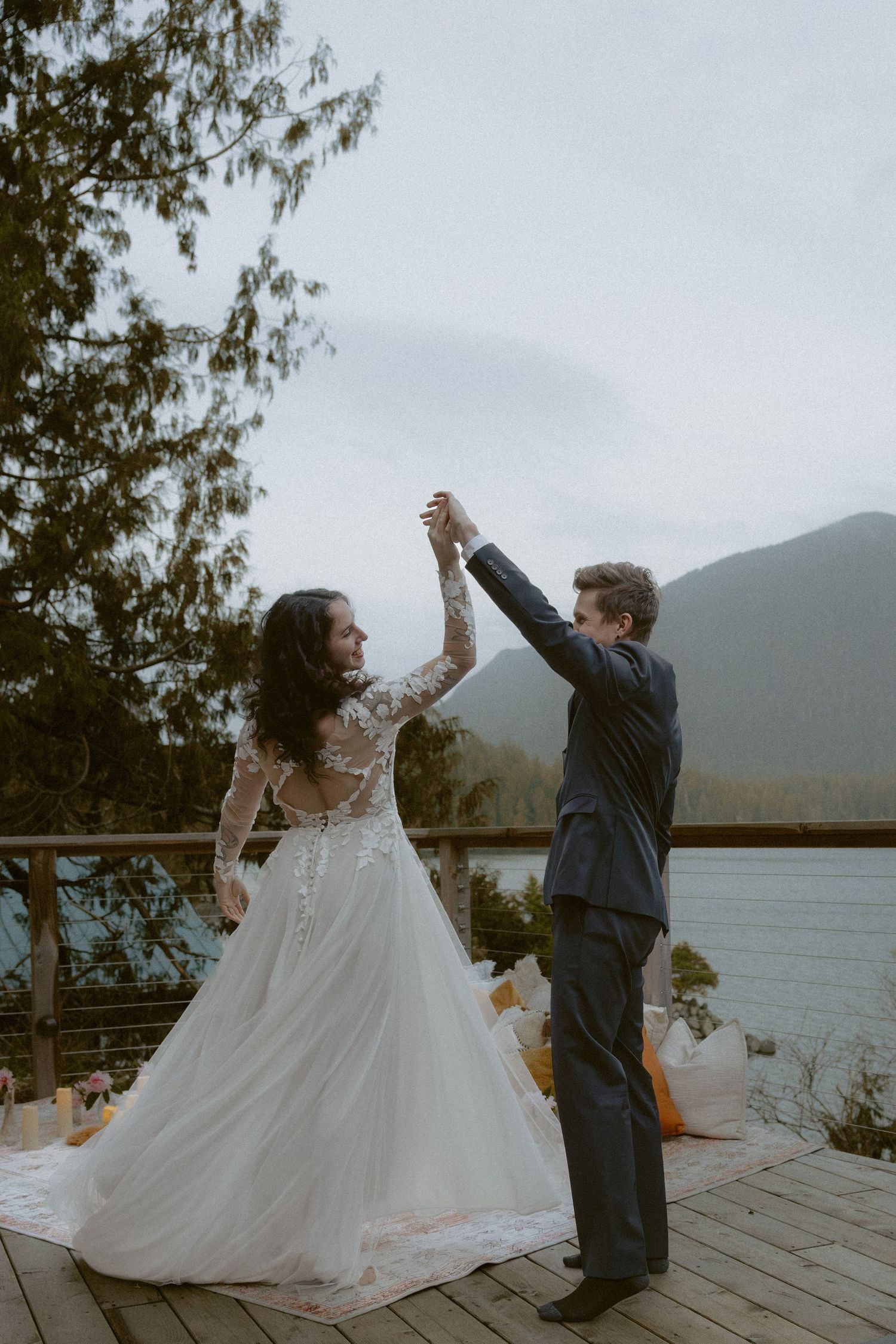 A couple dances joyfully on a wooden deck overlooking a misty mountain lake with evergreen trees in the background.