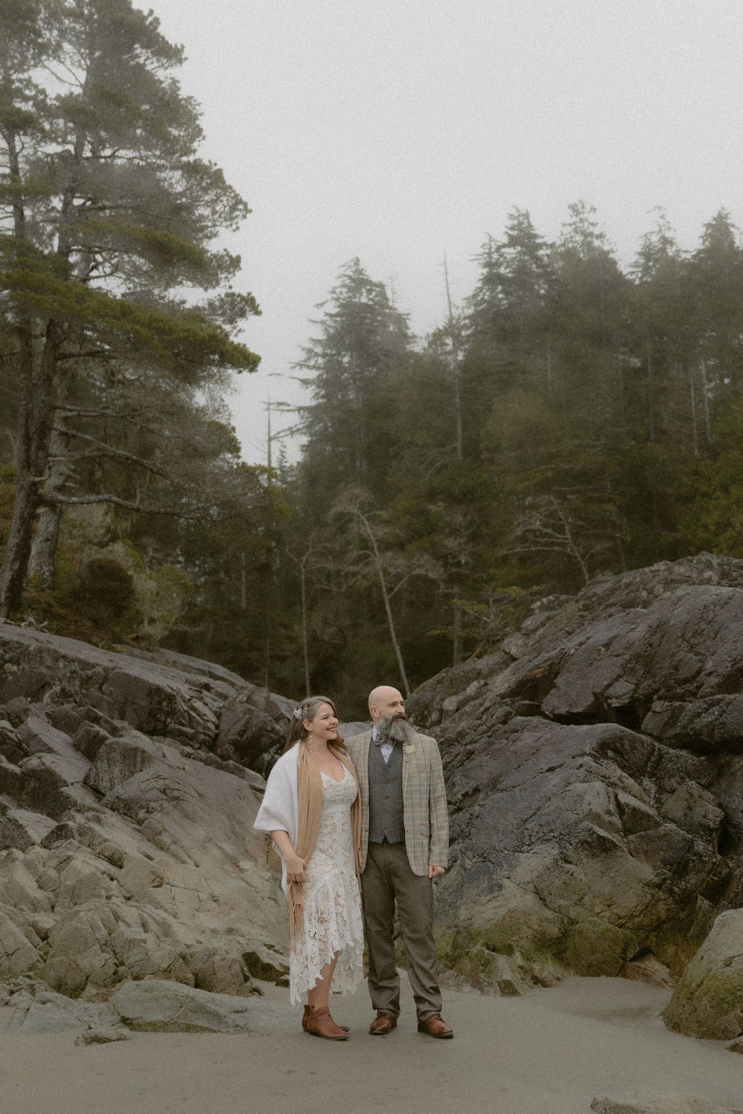 Couple standing together on a rocky beach with dramatic cliffs and evergreen forest in the background.
