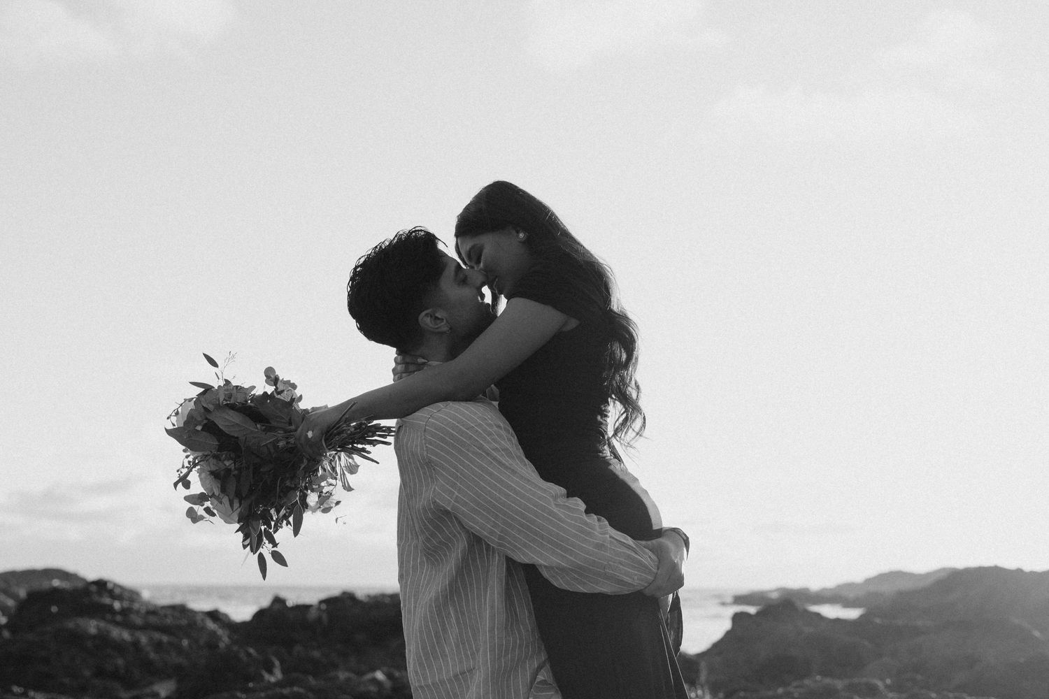Black and white photo of a man lifting his partner for a kiss while holding a bouquet outdoors.