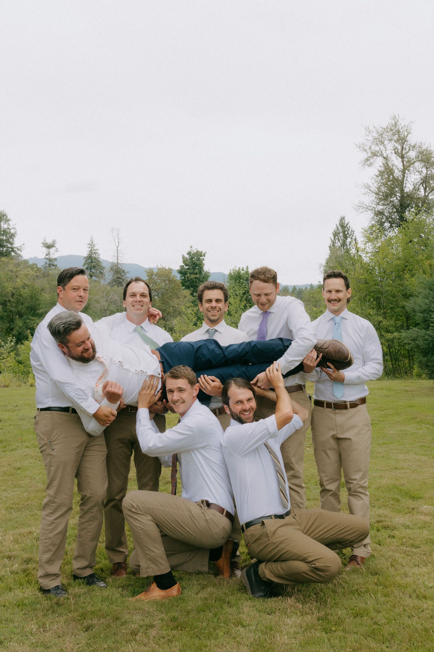 Groomsmen lifting groom horizontally in outdoor wedding photo on green lawn with trees in background.