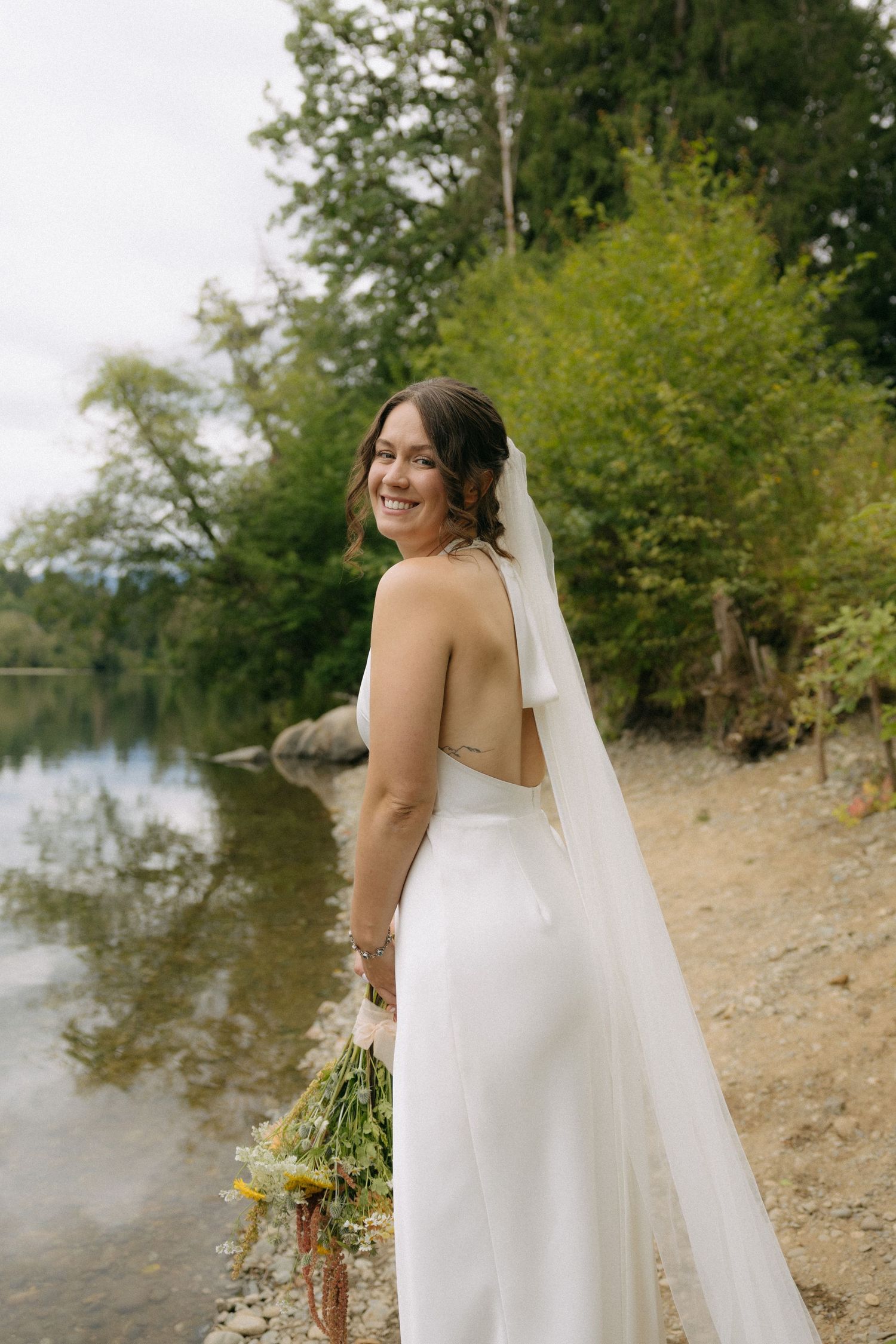 Smiling bride in backless white wedding gown and veil holds wildflowers beside a serene lake surrounded by trees.
