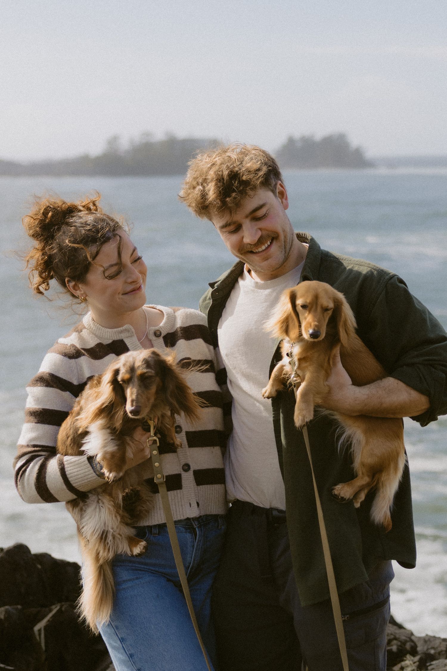 Happy couple laughing while holding two adorable golden dachshund puppies on a scenic rocky coastline.