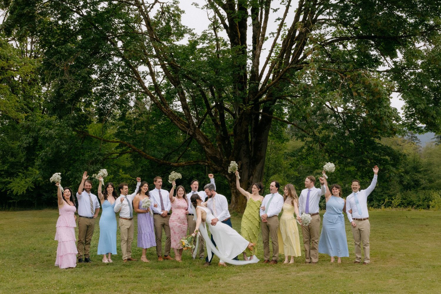 Wedding party celebrating outdoors on green lawn, bridesmaids in pastel dresses, groomsmen in khaki, bride and groom in center.