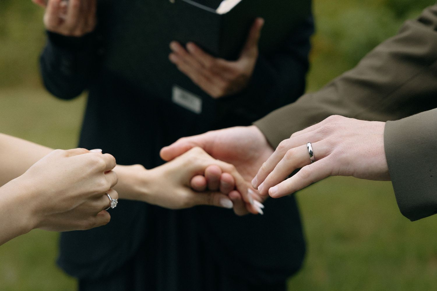 Couple exchanging rings during an outdoor wedding ceremony with an officiant holding a book.