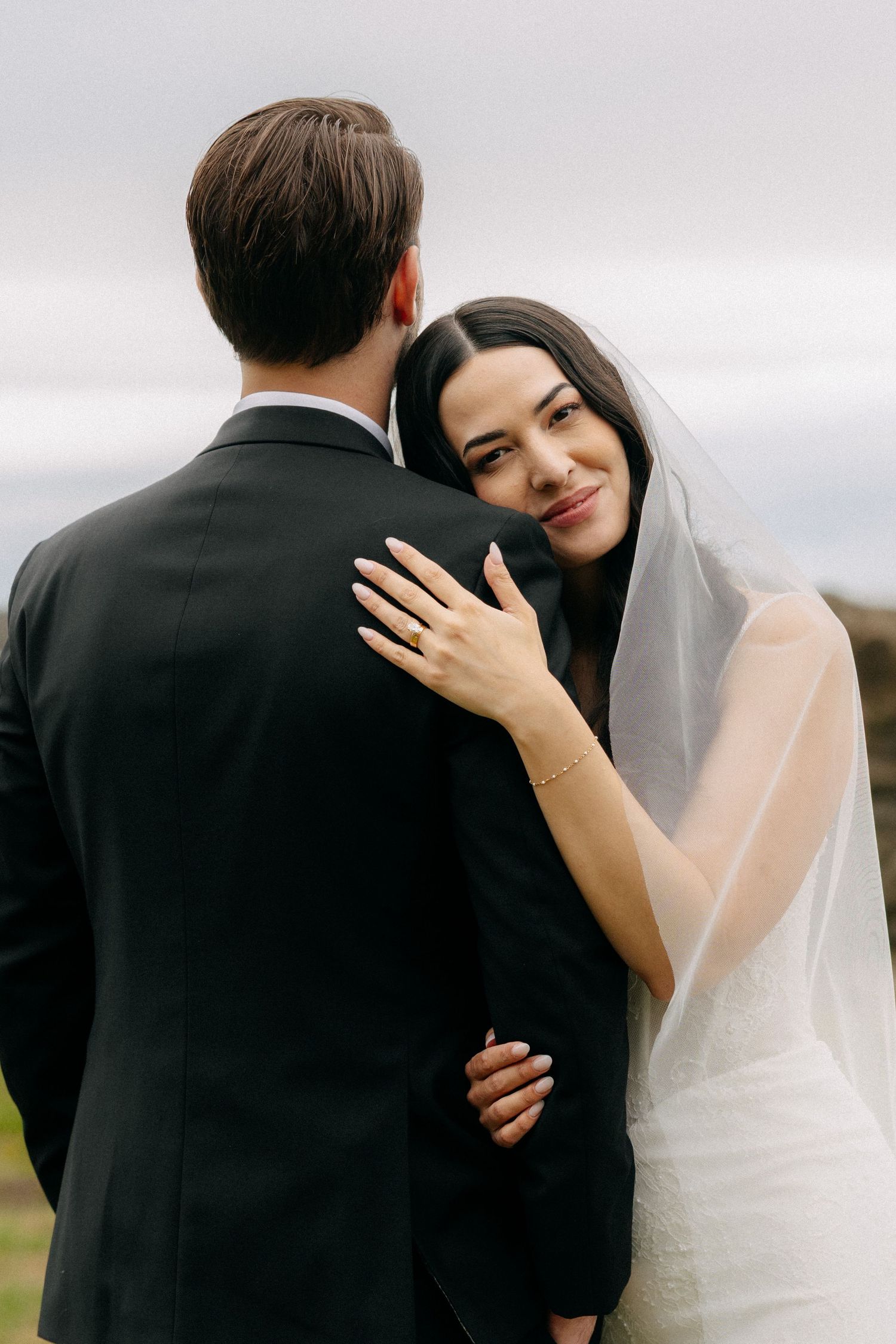 Bride in white gown and veil embraces groom in black suit outdoors, smiling over his shoulder.