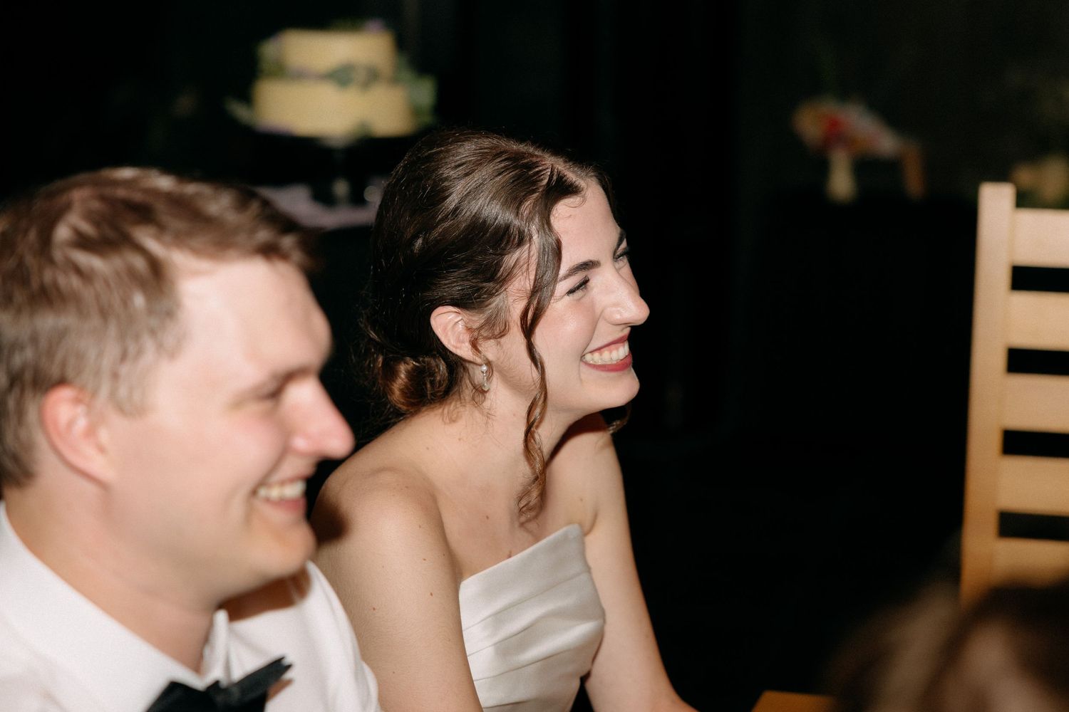 Bride and groom laughing joyfully at wedding reception, with wedding cake visible in background.