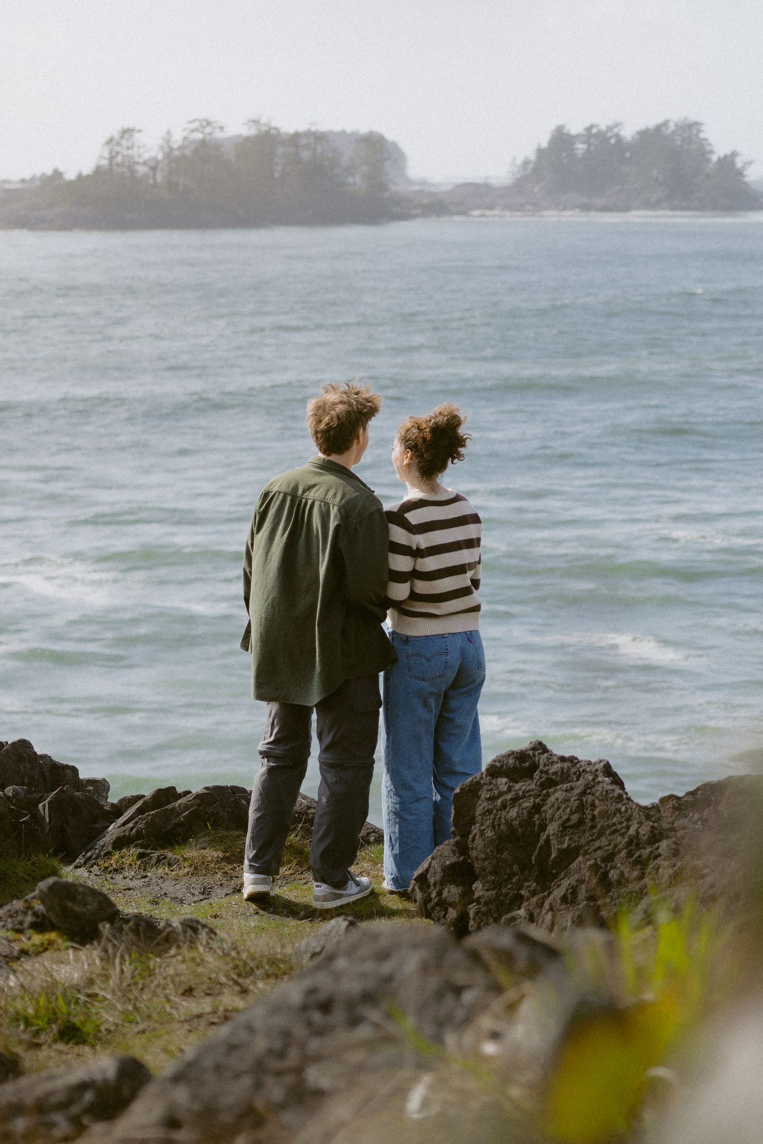 Couple standing together on rocky coastal cliffs overlooking a misty ocean with forested islands in the distance.