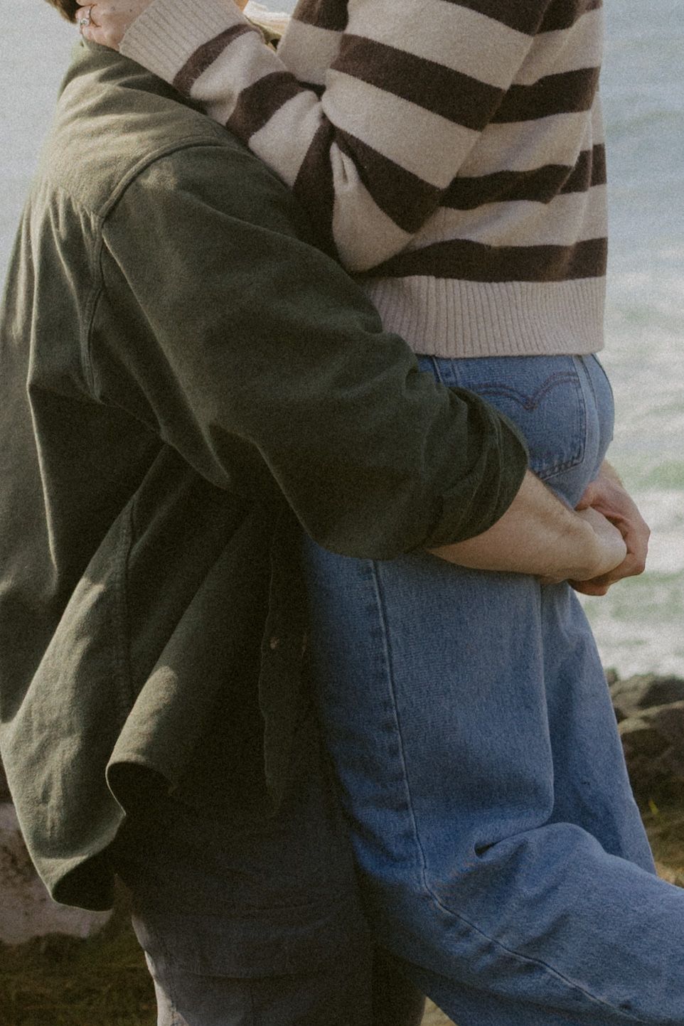 Close-up of a couple embracing, wearing a green jacket and striped sweater with blue jeans by the sea.