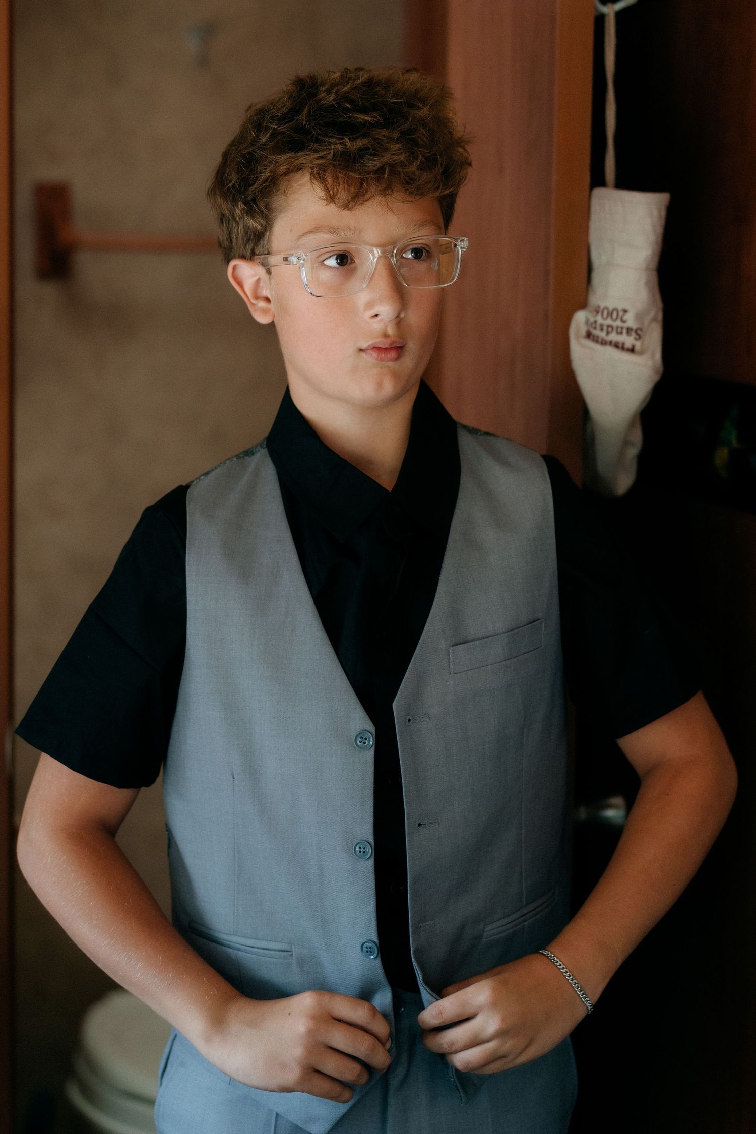 Young boy in glasses and gray vest over black shirt buttoning up, posing indoors for a formal occasion.