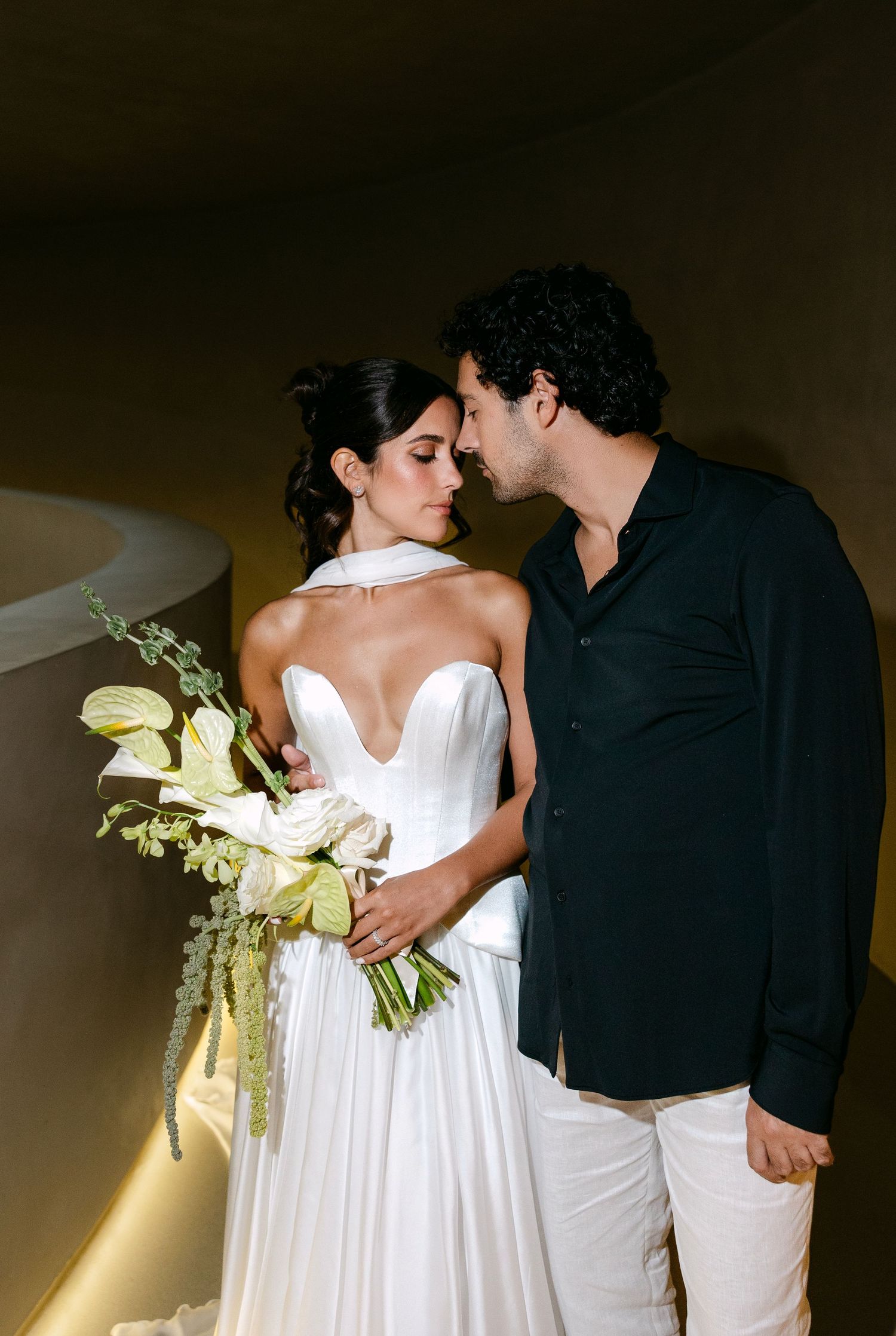Bride in strapless white gown with halter detail and groom in black shirt share intimate moment indoors.