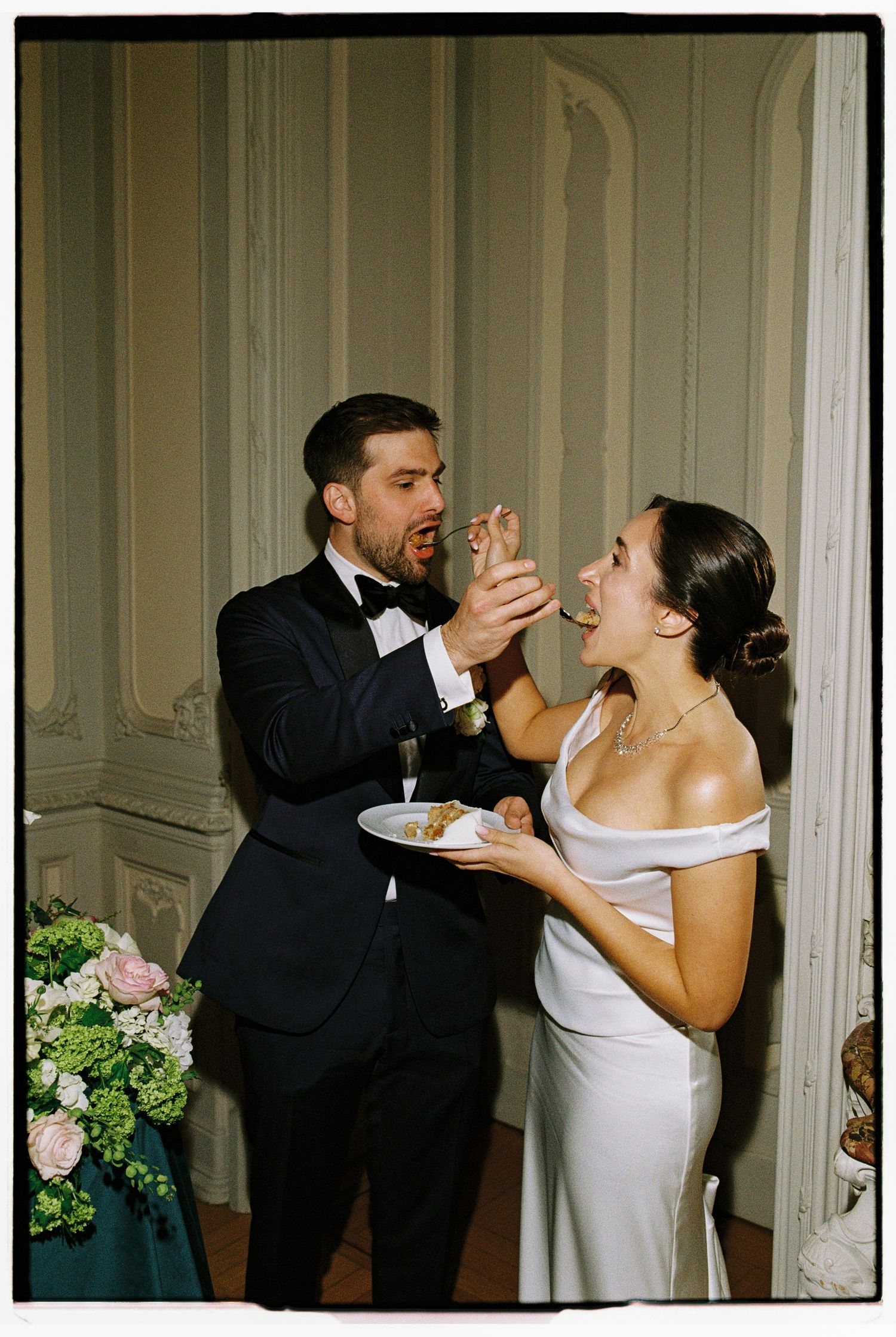 Bride and groom feeding each other wedding cake, wearing white gown and black tuxedo at elegant reception venue.