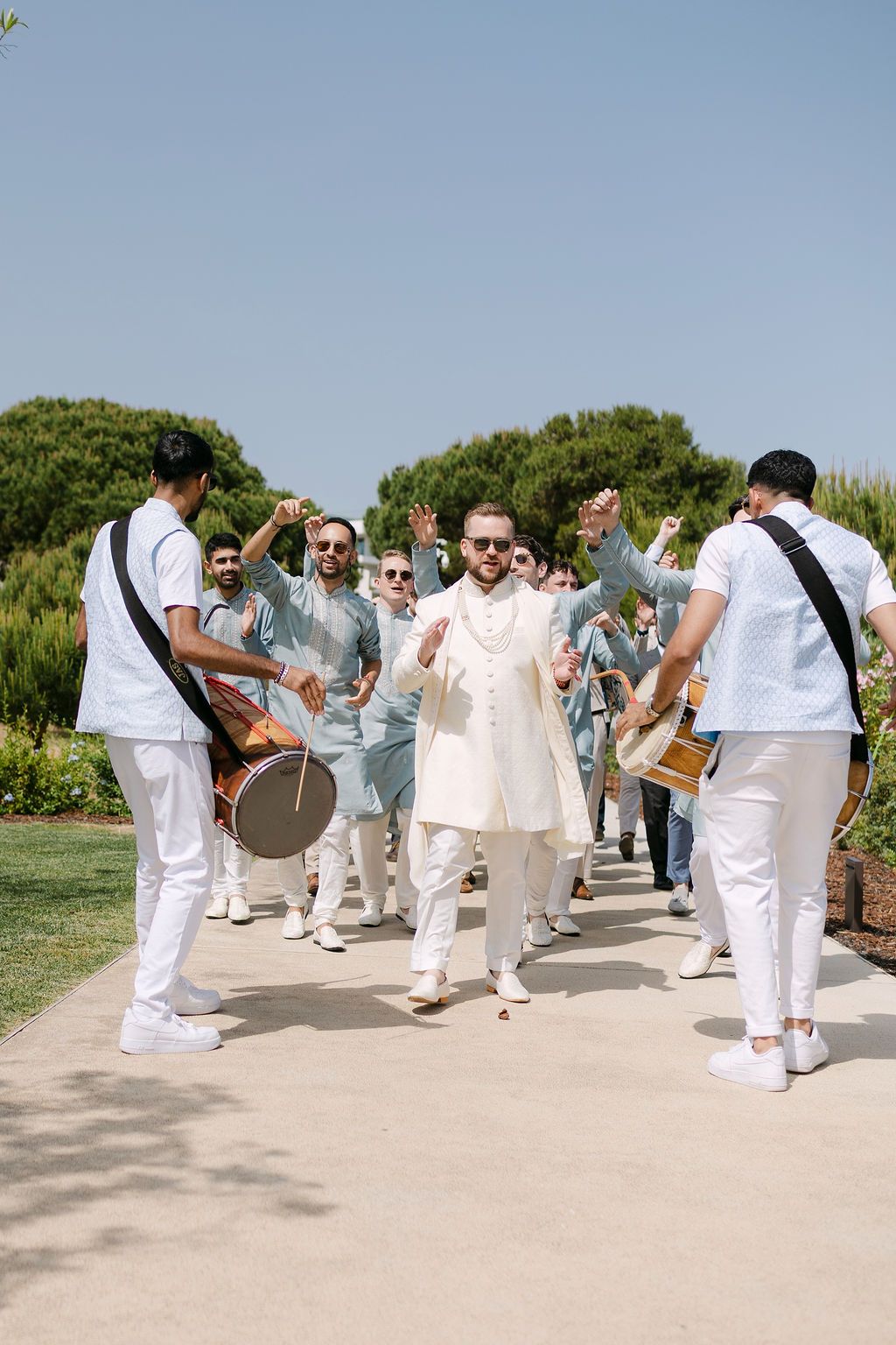 Wedding celebration procession with people dressed in white dancing and playing drums on an outdoor path.