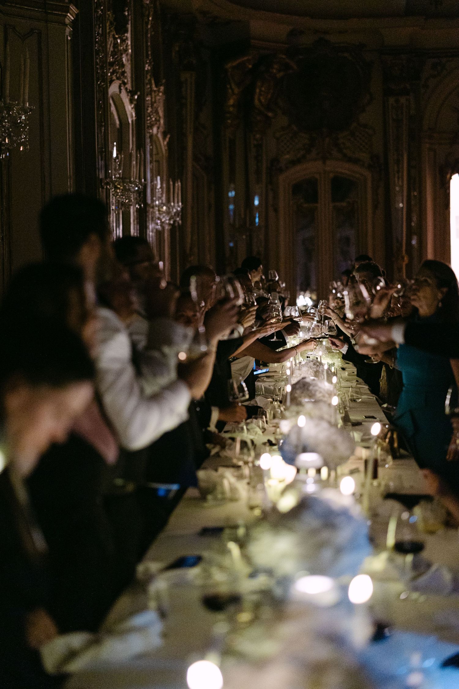 Wedding guests raise champagne glasses for toast in grand candlelit baroque dining hall.