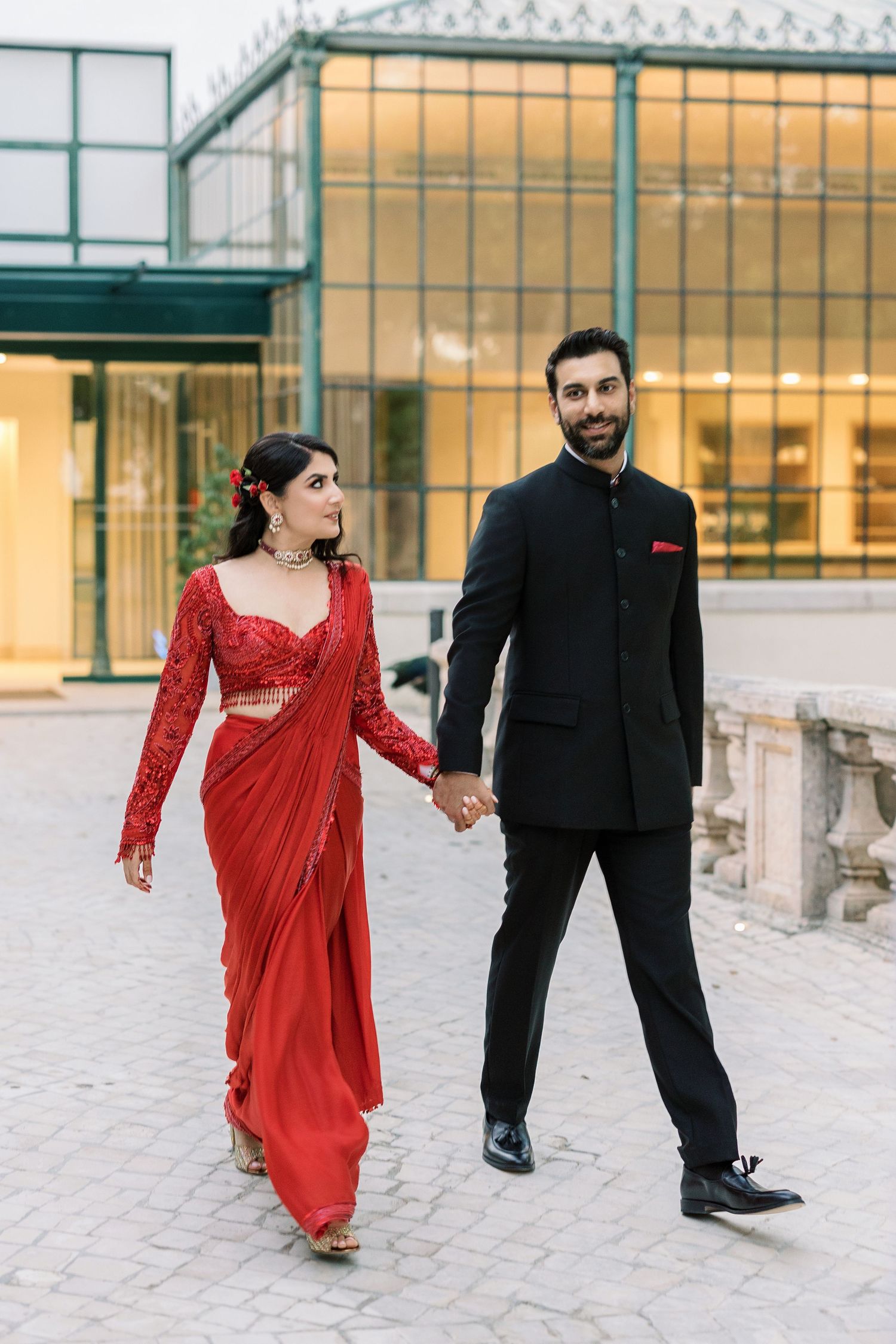 Couple walking outdoors in formal wear, with woman in red sari and man in black suit.