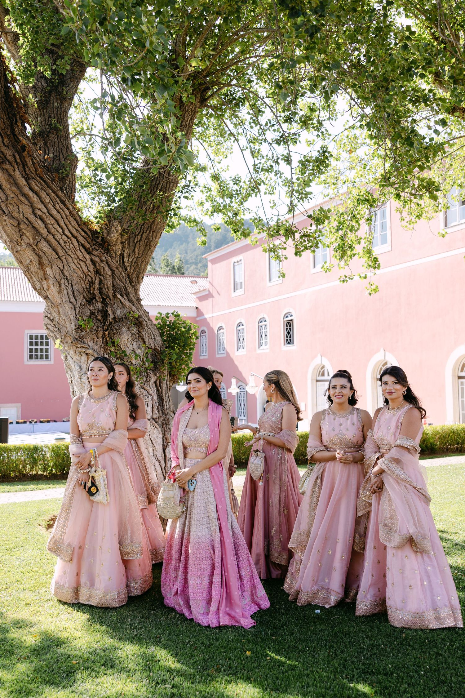 Bridesmaids in pink and rose-colored lehengas pose under large tree at elegant venue.