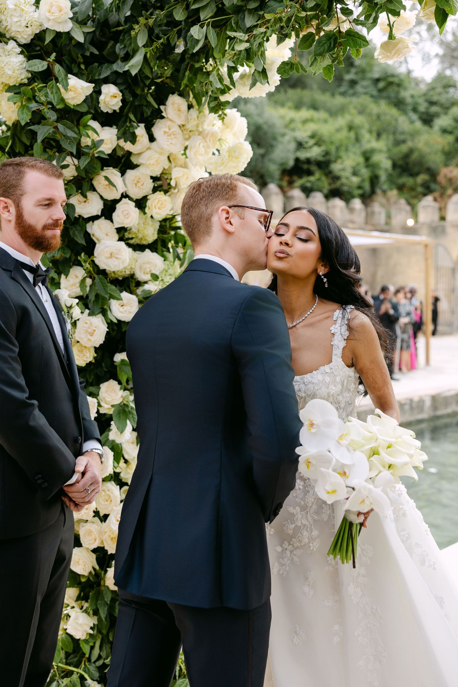 A romantic kiss under a floral arch with white roses during an outdoor wedding ceremony.