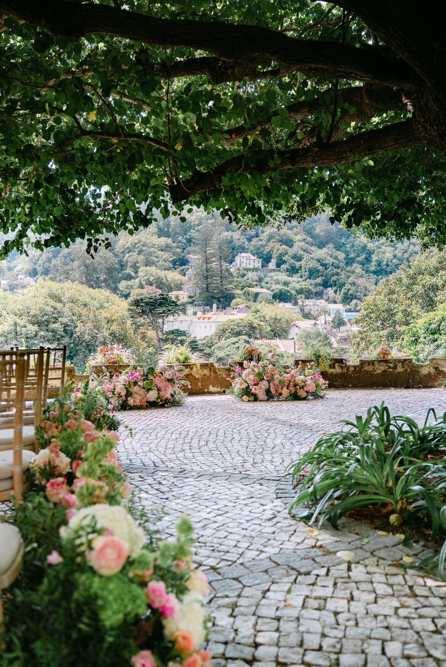 Stone paved outdoor ceremony space decorated with pink floral arrangements and wooden chairs.