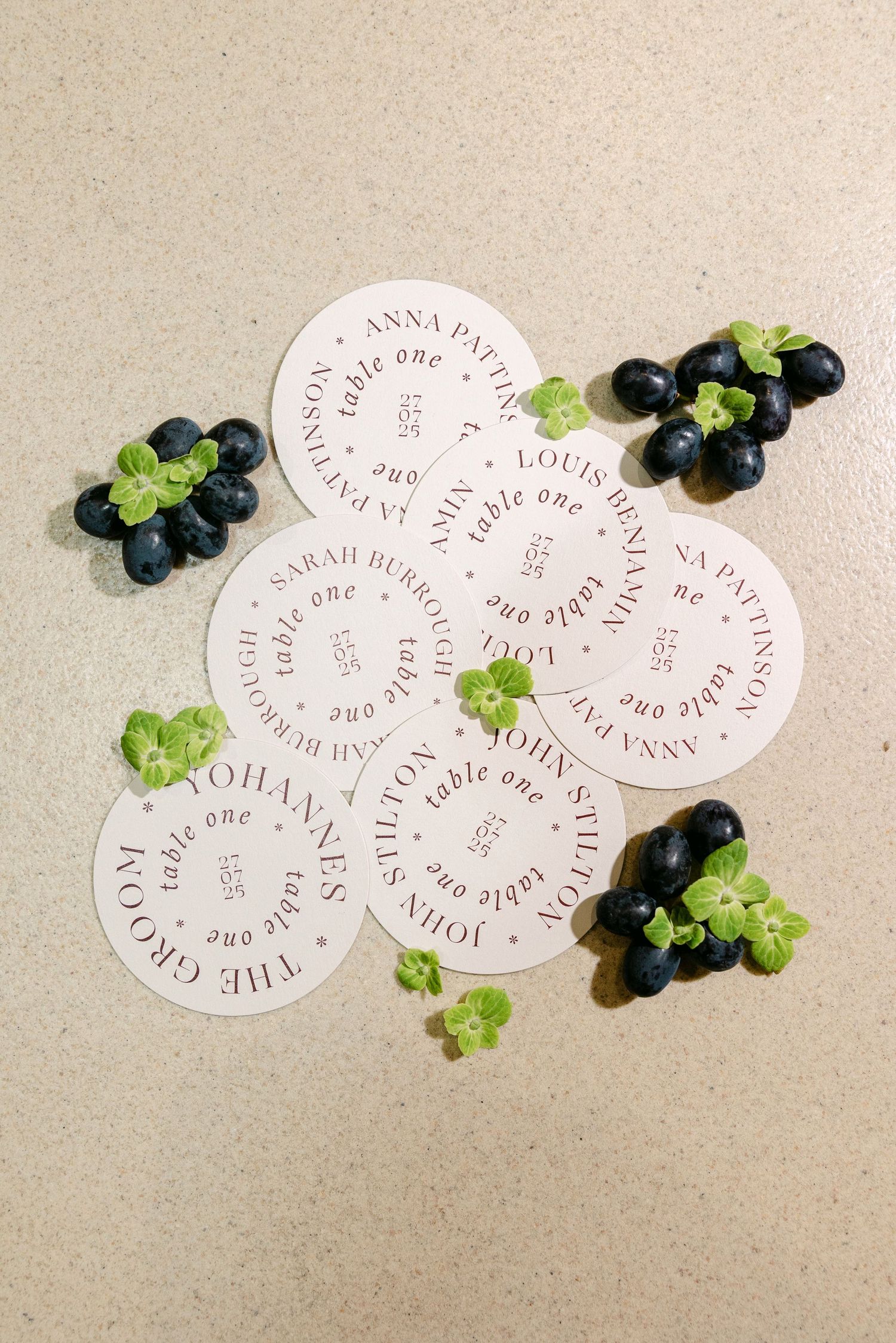 Circular wedding place cards with guest names arranged with fresh grapes and green leaves on a neutral surface.