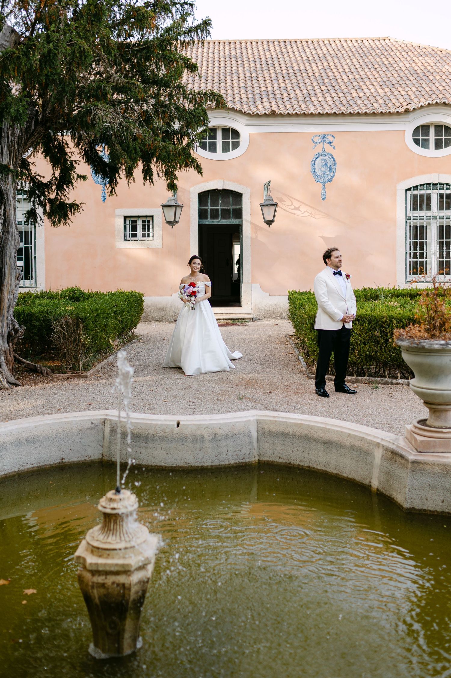 Wedding couple poses near a decorative fountain outside a Mediterranean-style building.