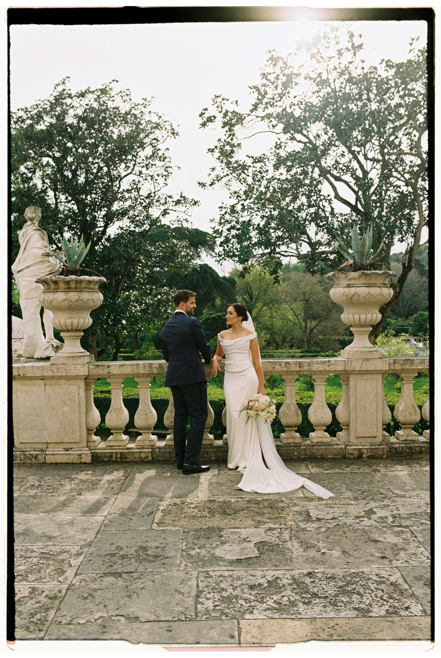 Bride and groom share intimate moment on stone terrace with classical balustrade and garden views.