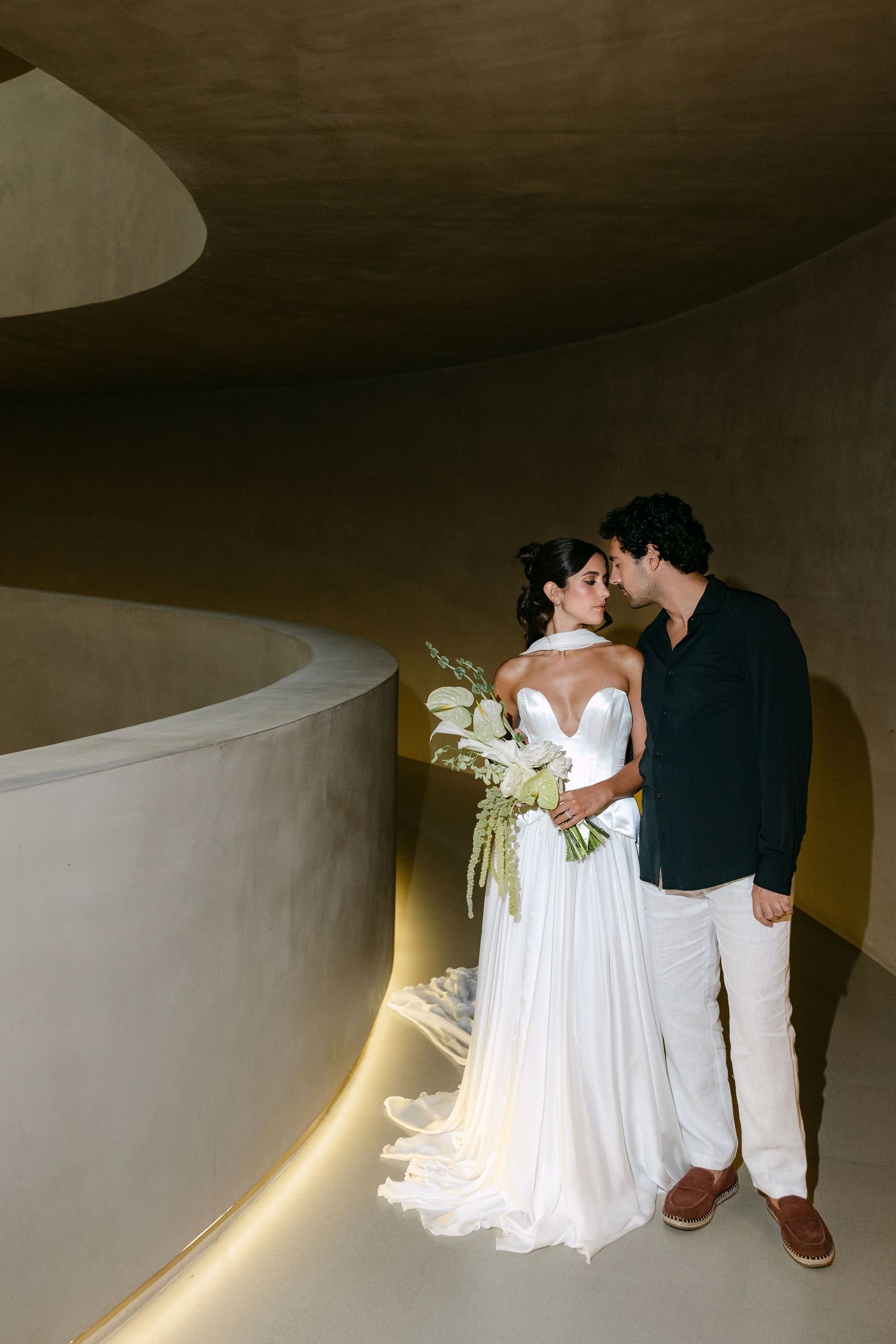 Bride and groom share a kiss in a curved architectural hallway, bride holding white floral bouquet in elegant strapless gown.