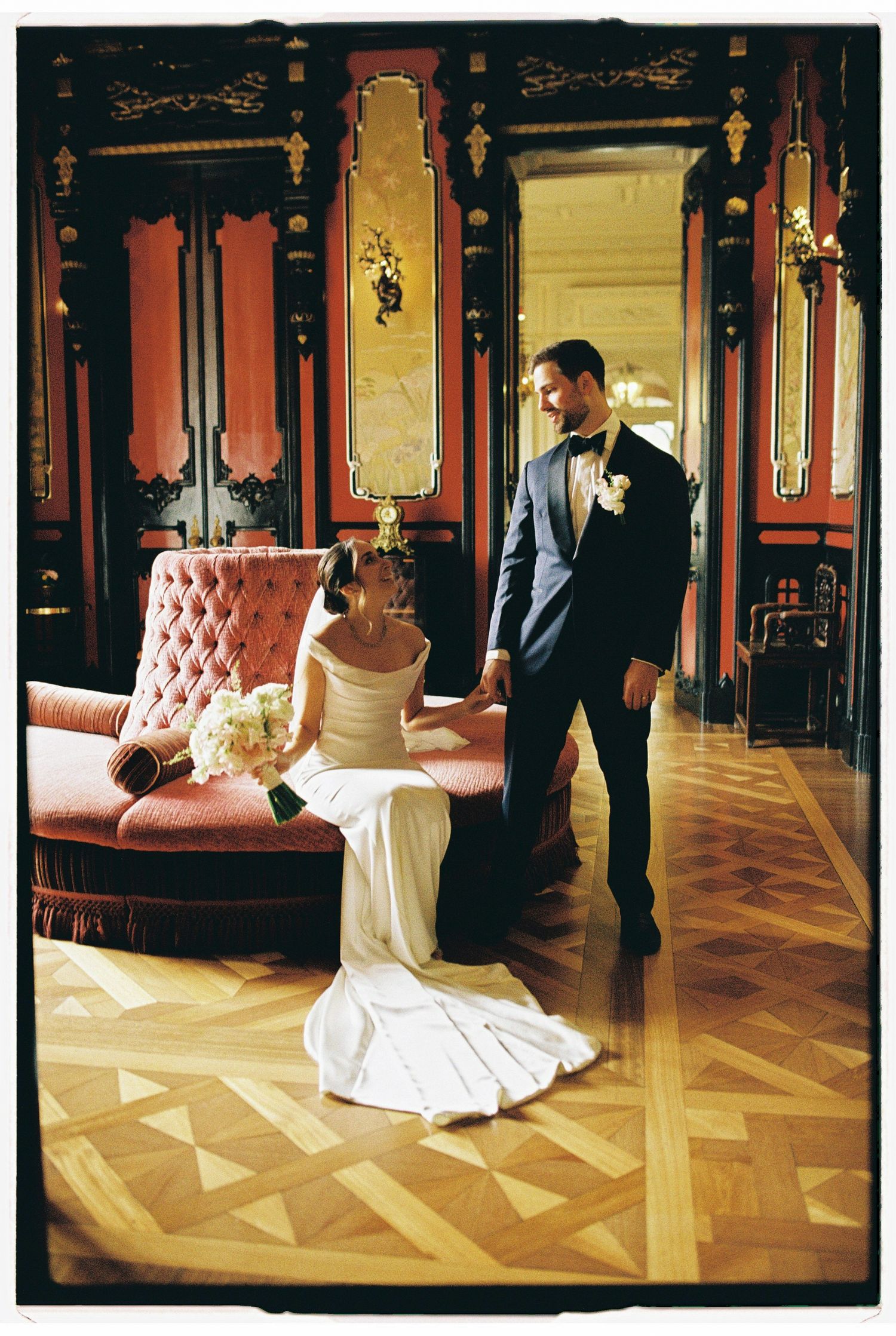 Bride in white gown sits on pink tufted sofa holding hands with groom in blue suit in ornate red ballroom.