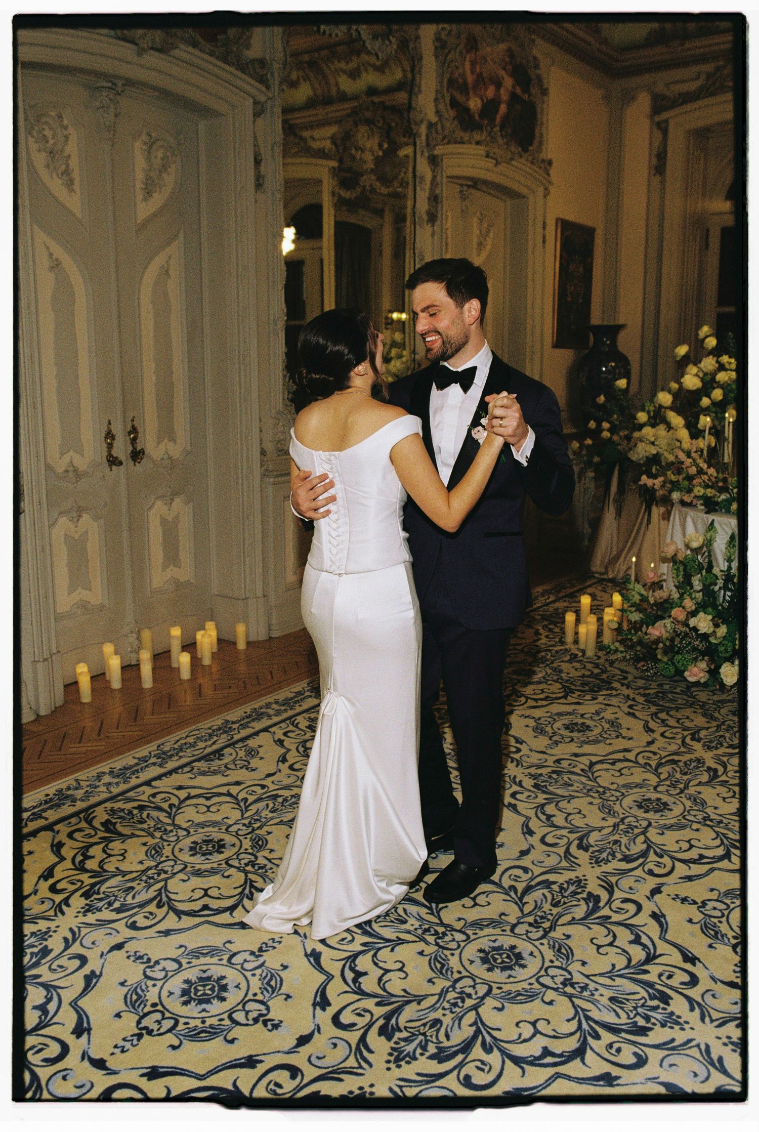 Bride and groom share their first dance in an elegant ballroom with ornate decor, candles, and a floral patterned carpet.
