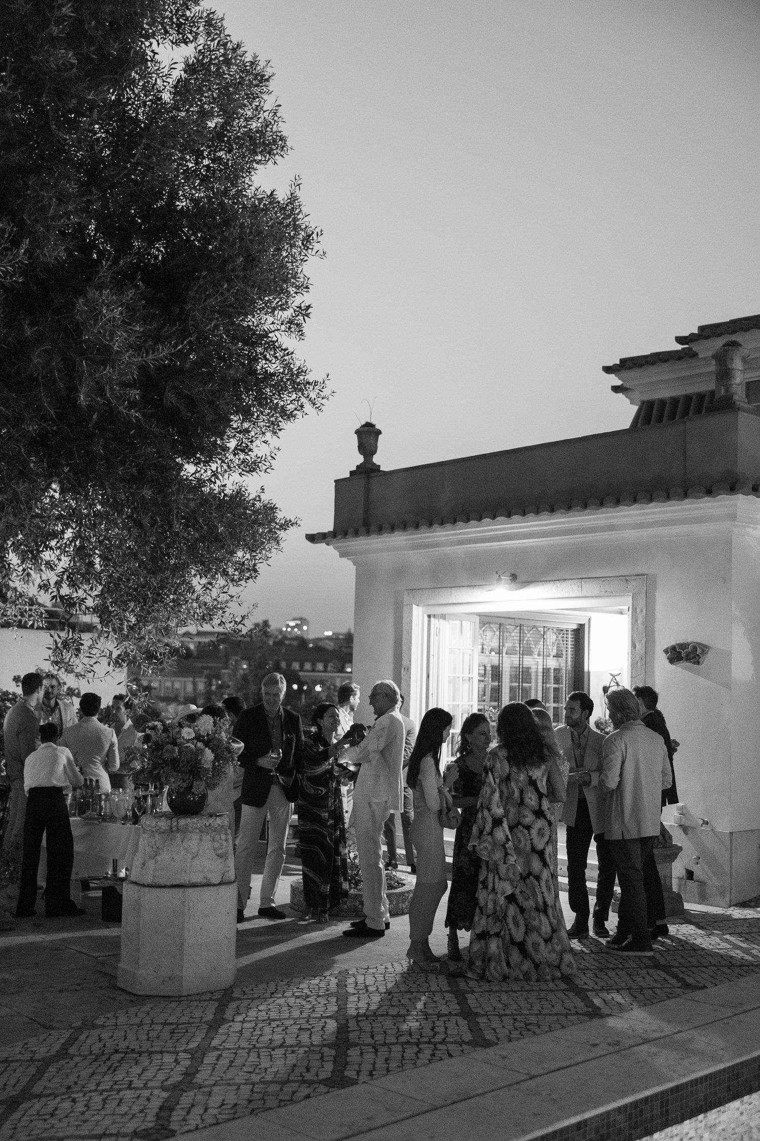 Black and white photo of elegantly dressed guests mingling outside a white villa entrance at dusk.