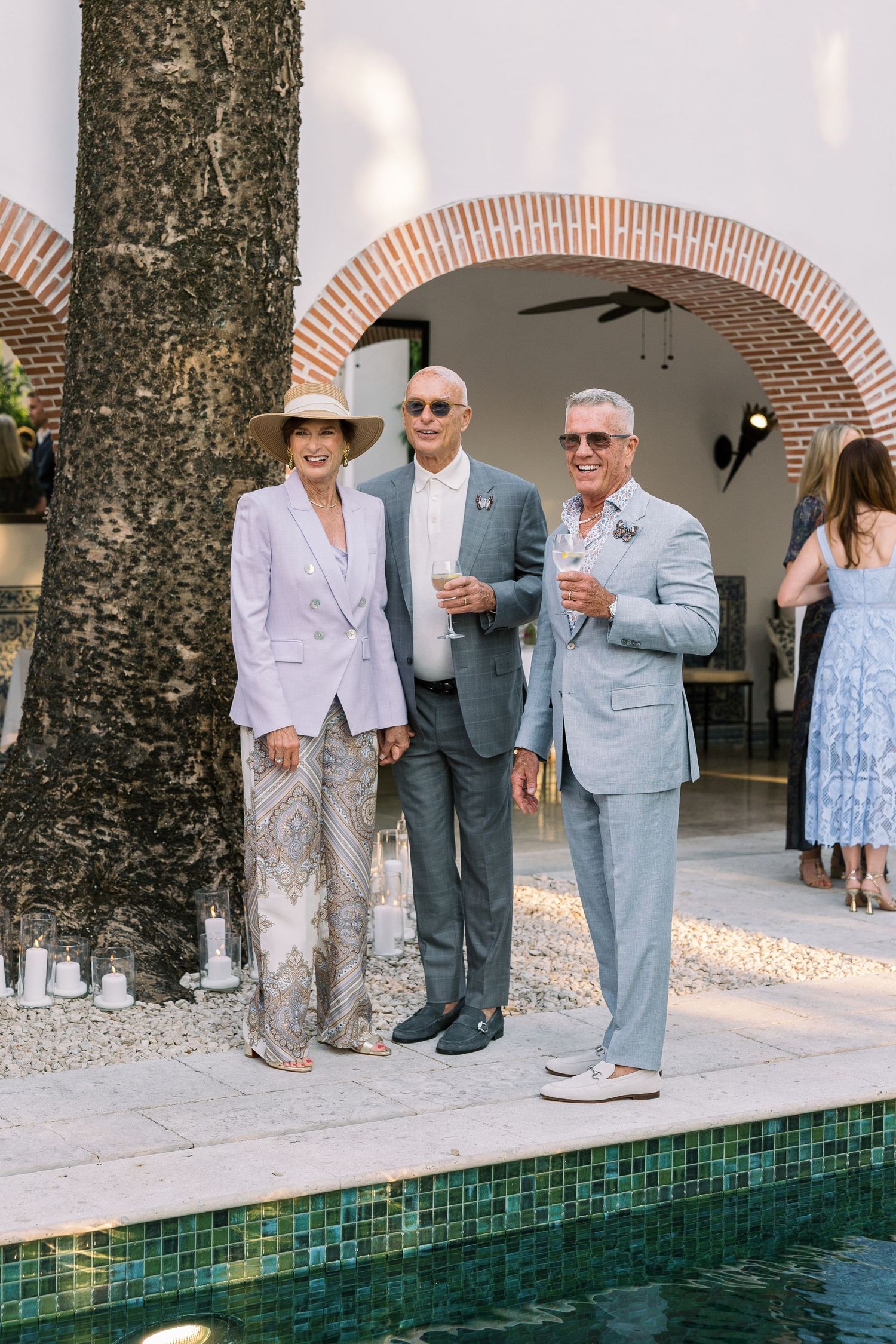 Three elegantly dressed guests pose poolside at an outdoor wedding reception near a brick archway.
