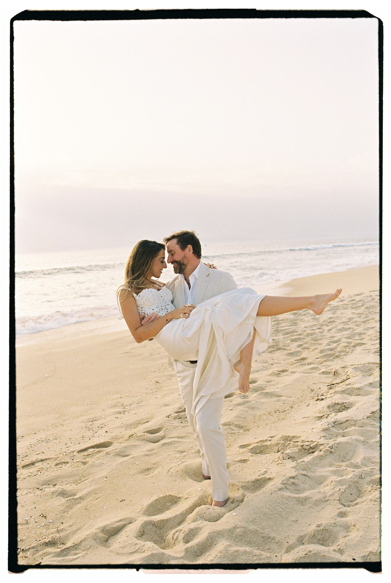 Couple in white embracing on a sandy beach at sunset, man lifting woman romantically.