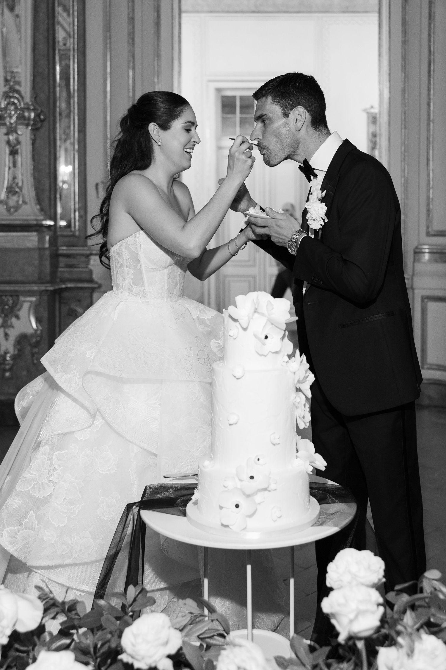 Bride feeds groom wedding cake slice beside white floral tiered cake in ornate room.