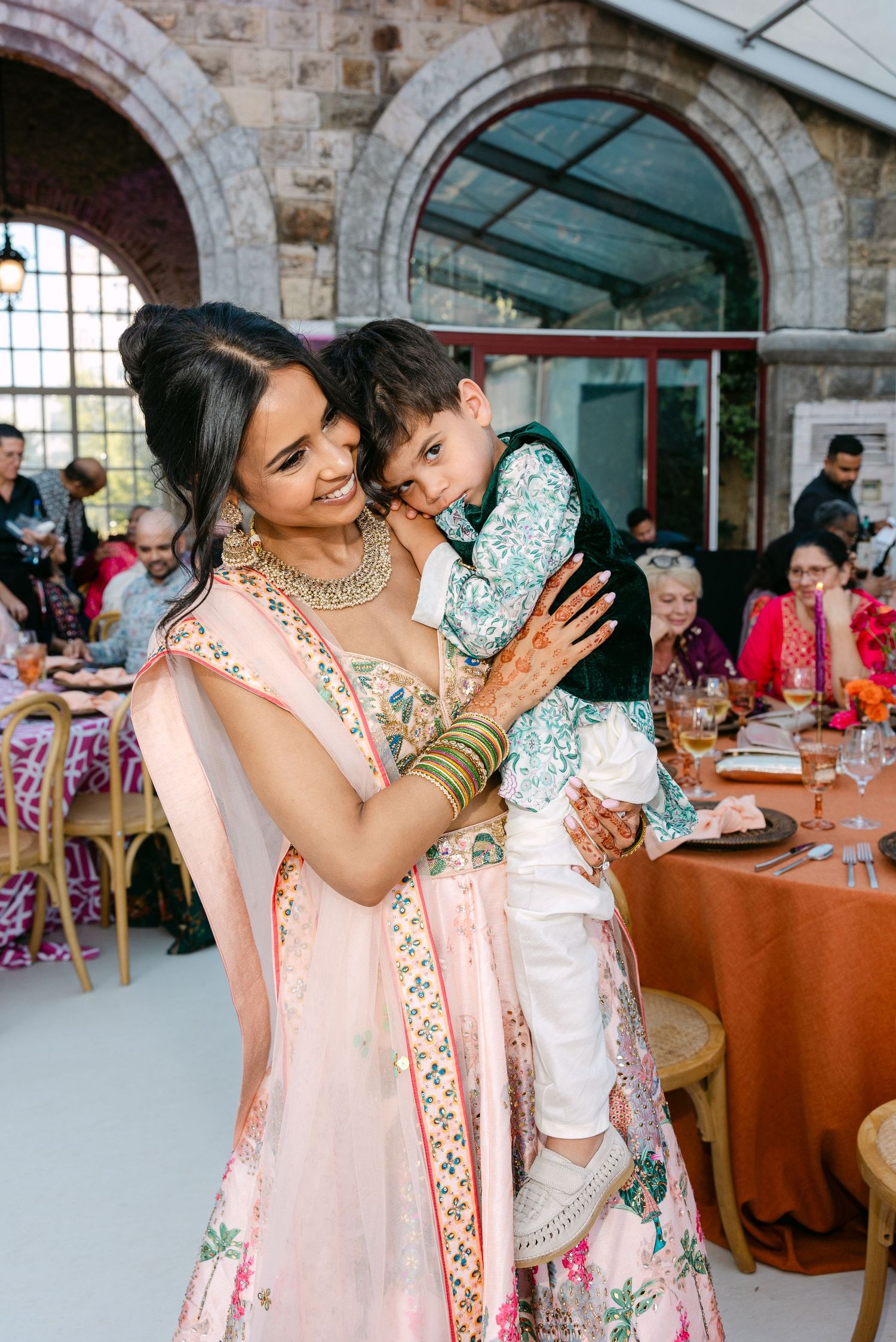 Wedding guests in colorful traditional Indian attire embracing and celebrating at an indoor venue.