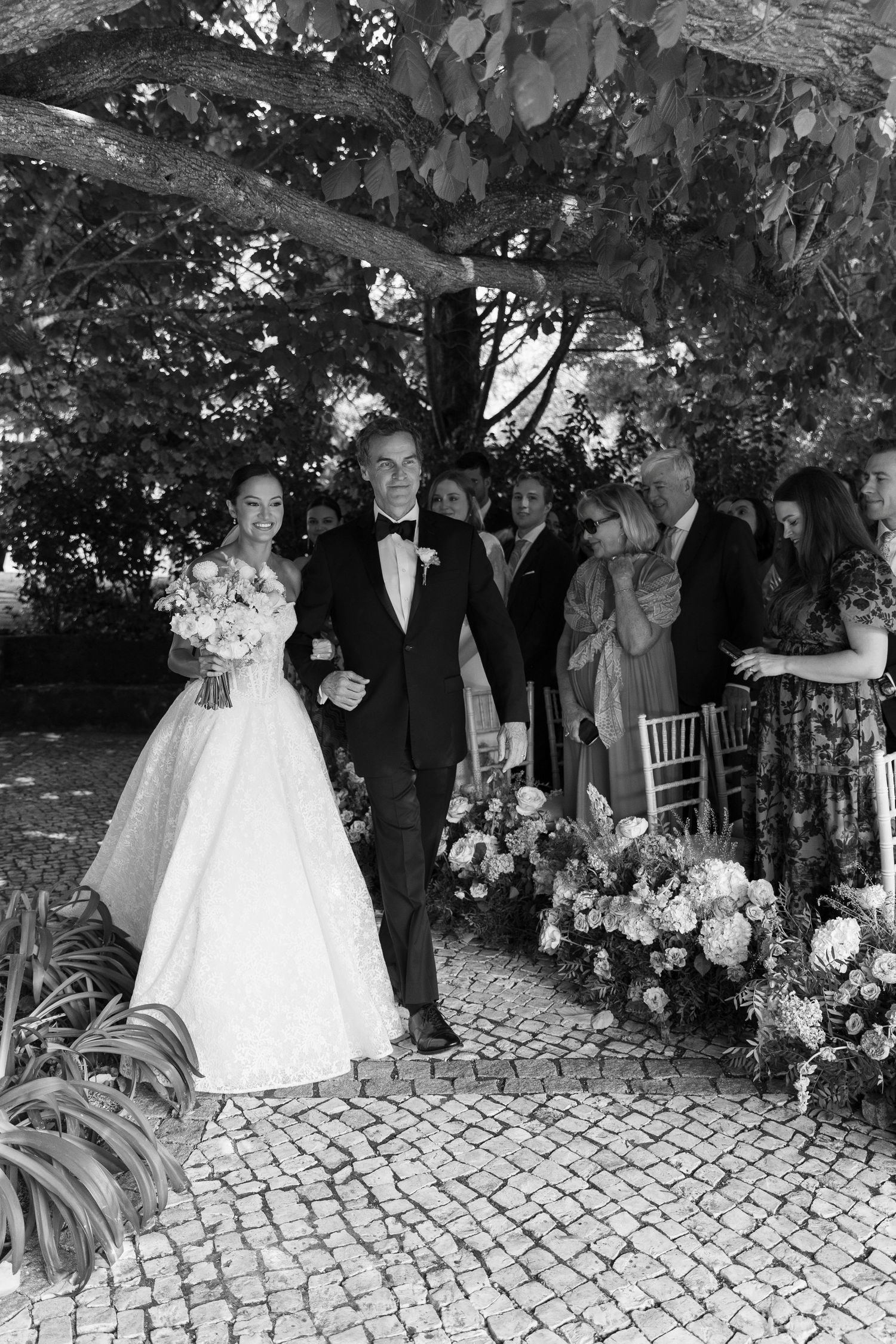 Black and white photo of wedding ceremony procession on cobblestone path under tree canopy.