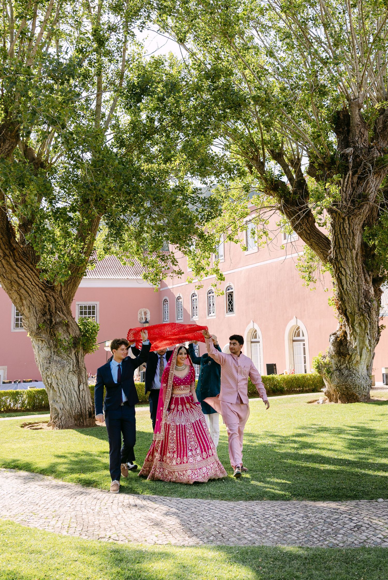 Wedding party walking on garden path beside pink building with trees.