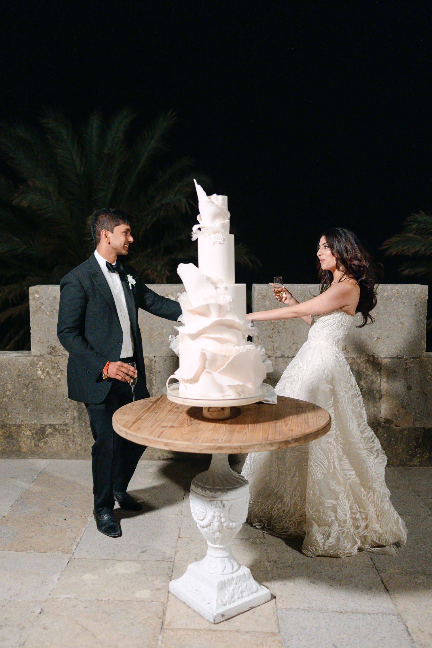 A couple admires their elegant white modern wedding cake on an ornate pedestal table outdoors.
