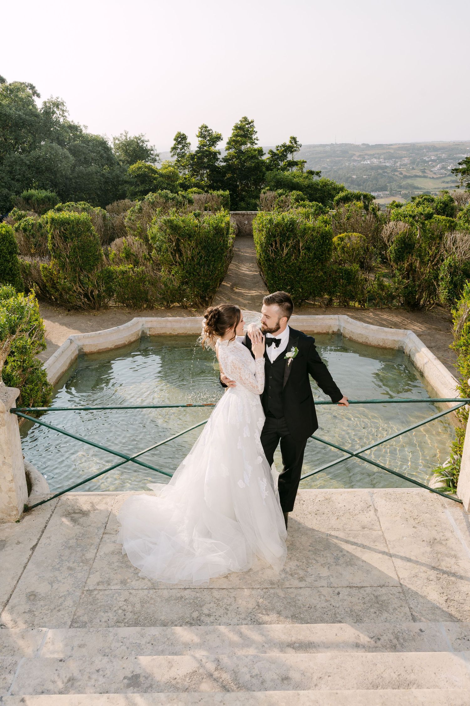 Wedding couple poses by stone balcony overlooking scenic mountain landscape.