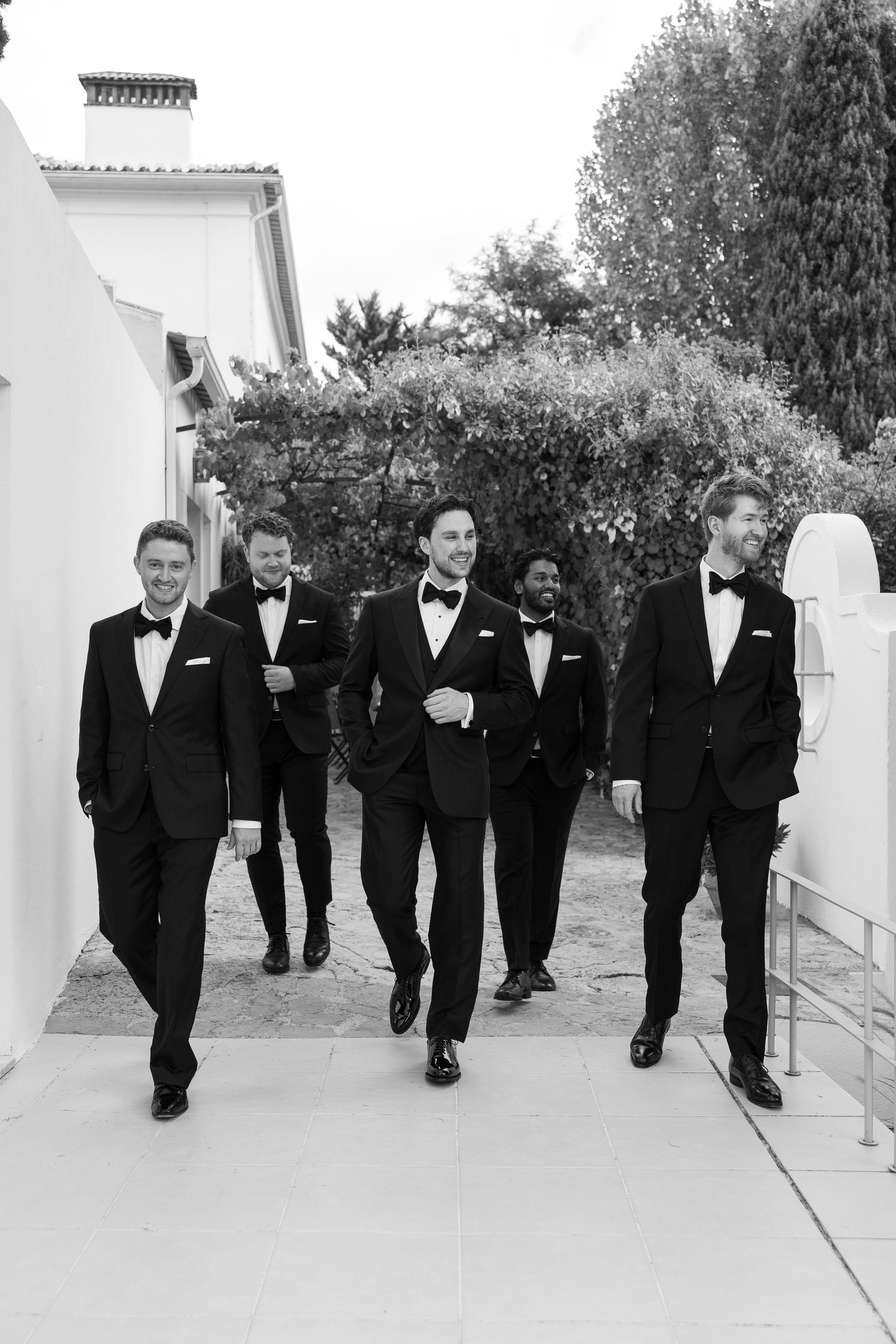 Five groomsmen in black tuxedos and bow ties walk confidently together outdoors in a classic black and white wedding photo.