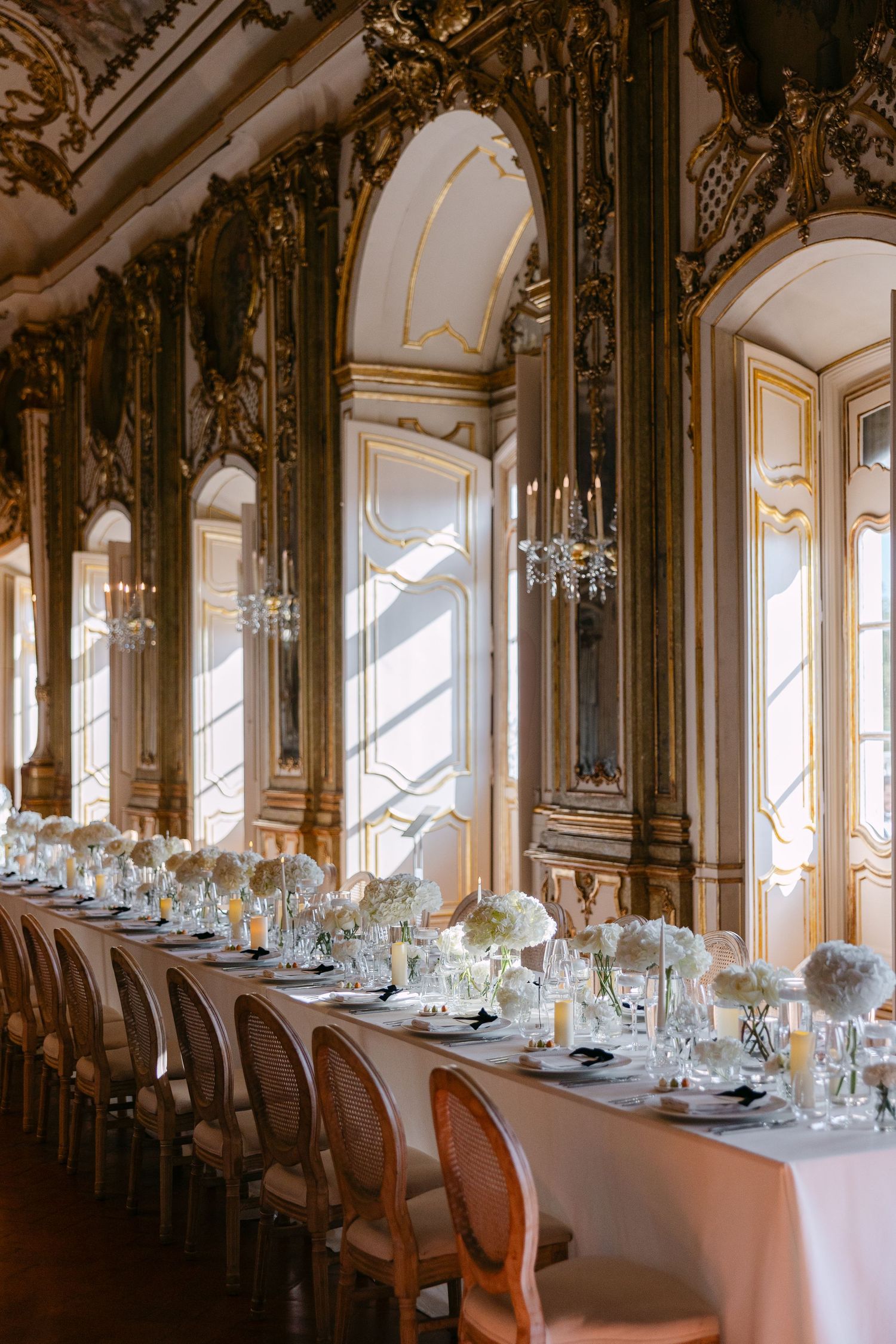 Elegant wedding reception in ornate baroque ballroom with long white table, floral centerpieces, and gilded architectural details.