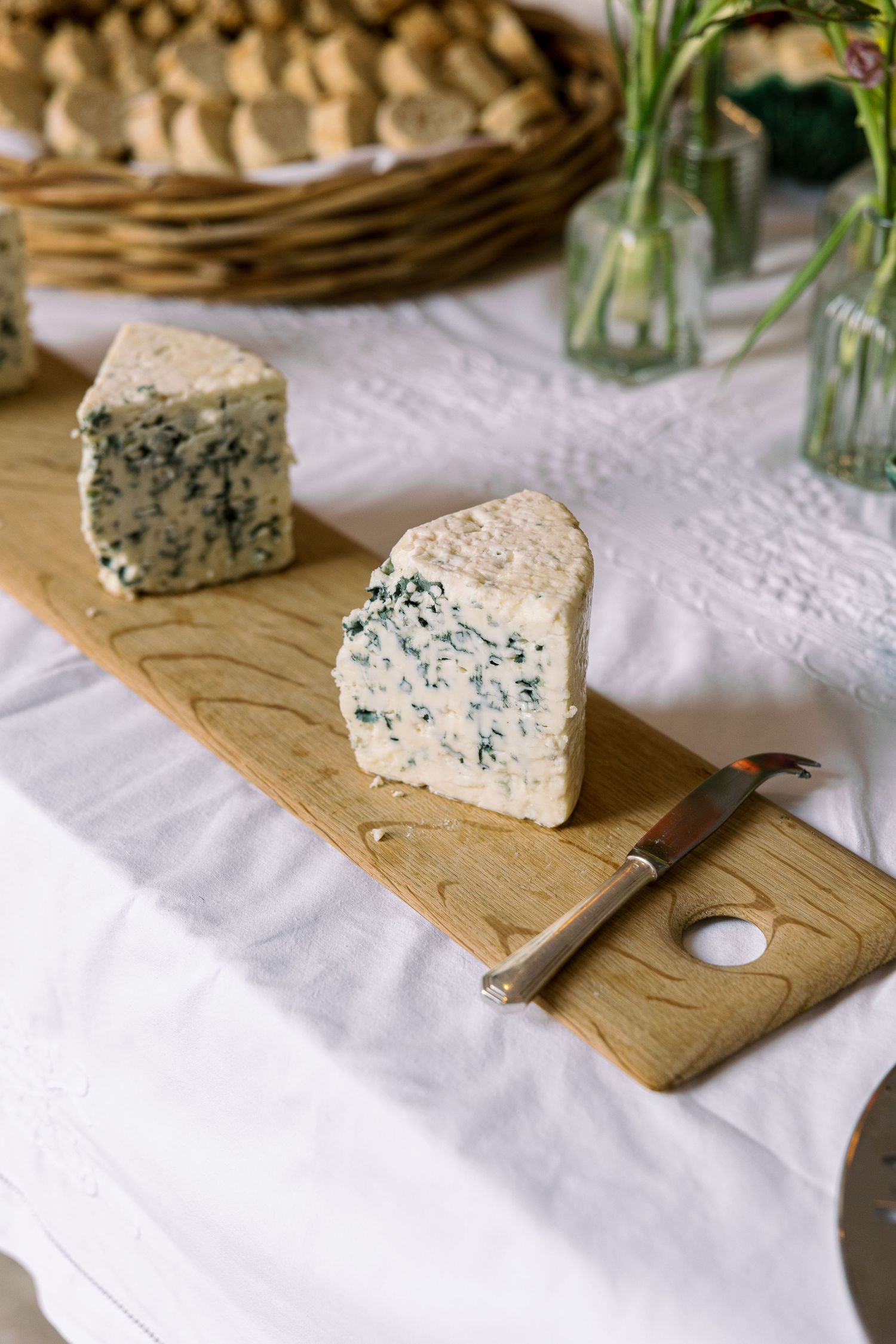Two wedges of blue cheese on a wooden cutting board with a cheese knife, surrounded by bread and flowers.