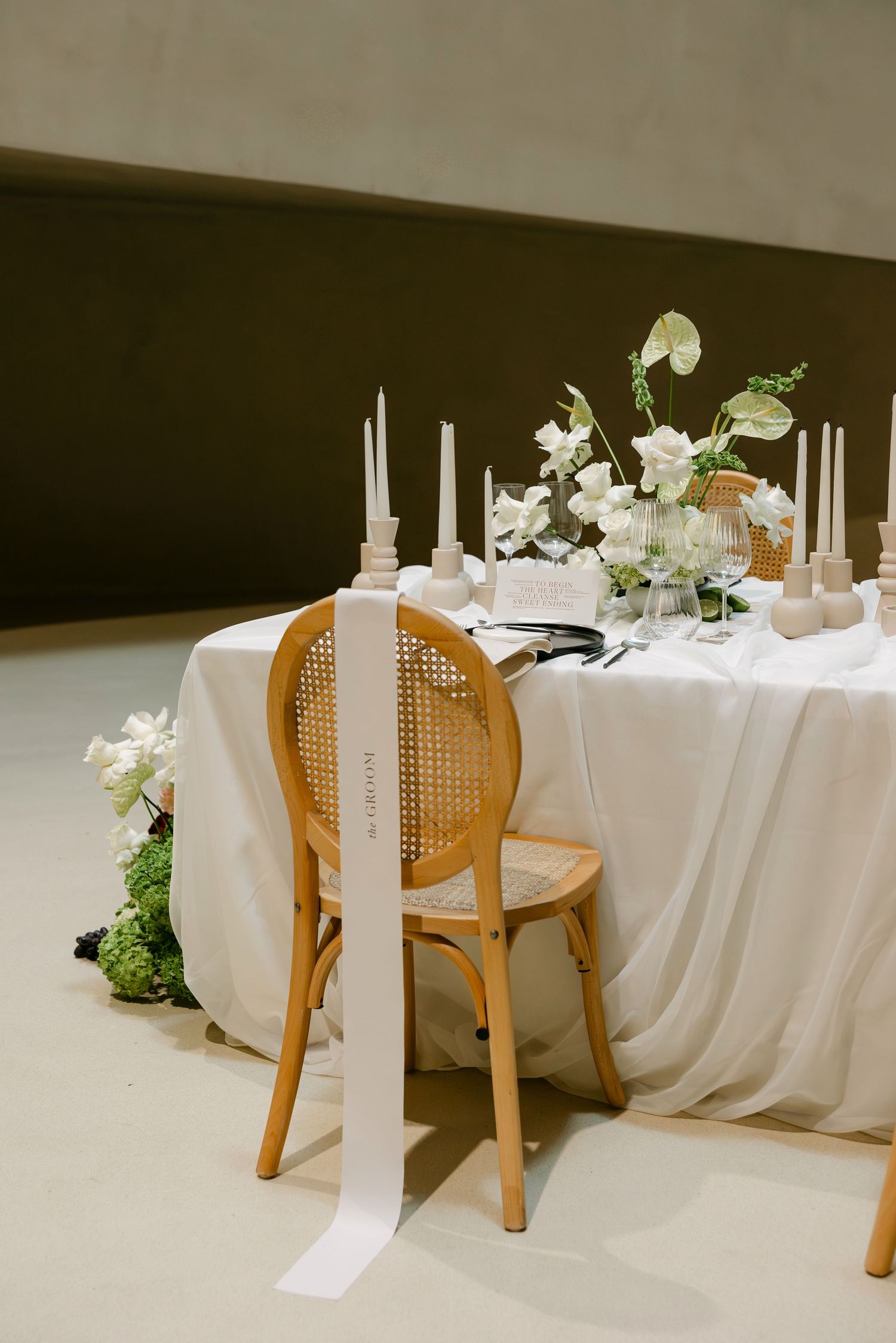 Rattan chair with white ribbon at elegant wedding sweetheart table with candles and white florals.
