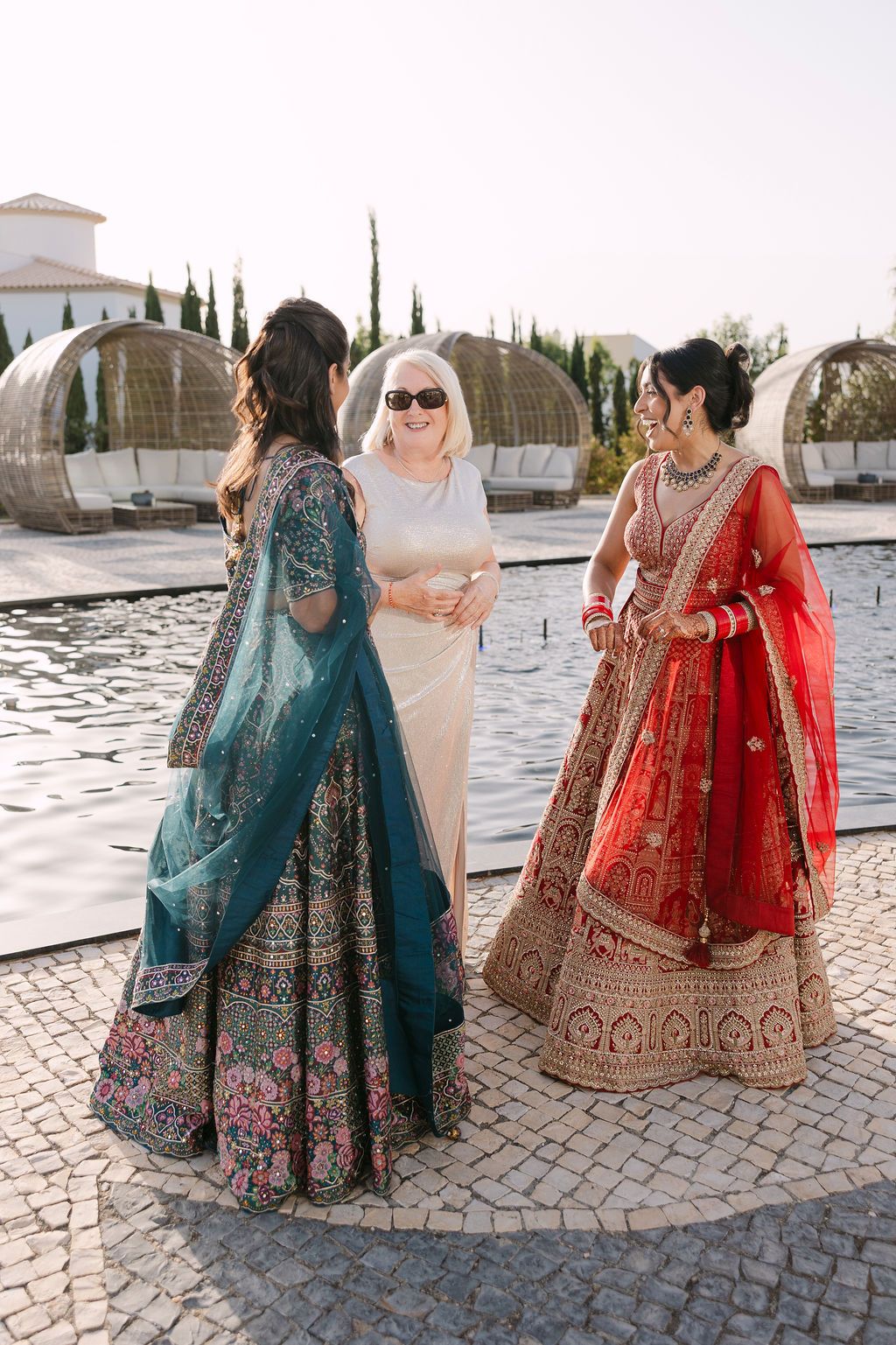 Traditional Indian wedding attire showcased in red and teal lehengas against Mediterranean architecture.