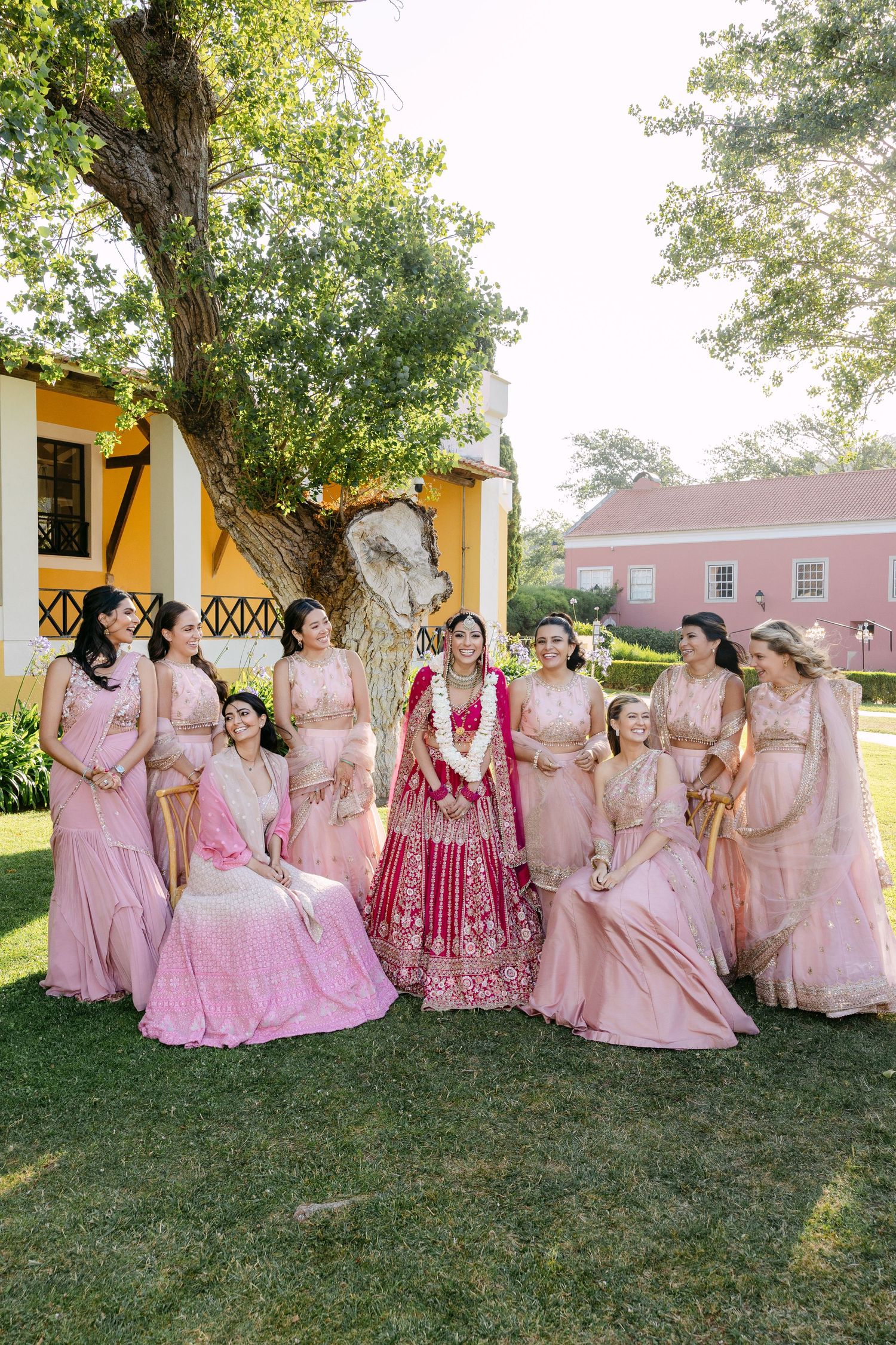 Wedding party in pink and red traditional Indian attire posing on grass near yellow building.