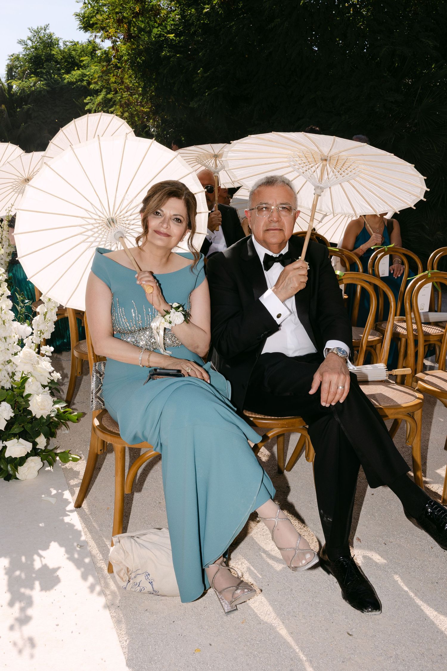 Wedding guests with white parasols seated on wooden chairs in formal attire.