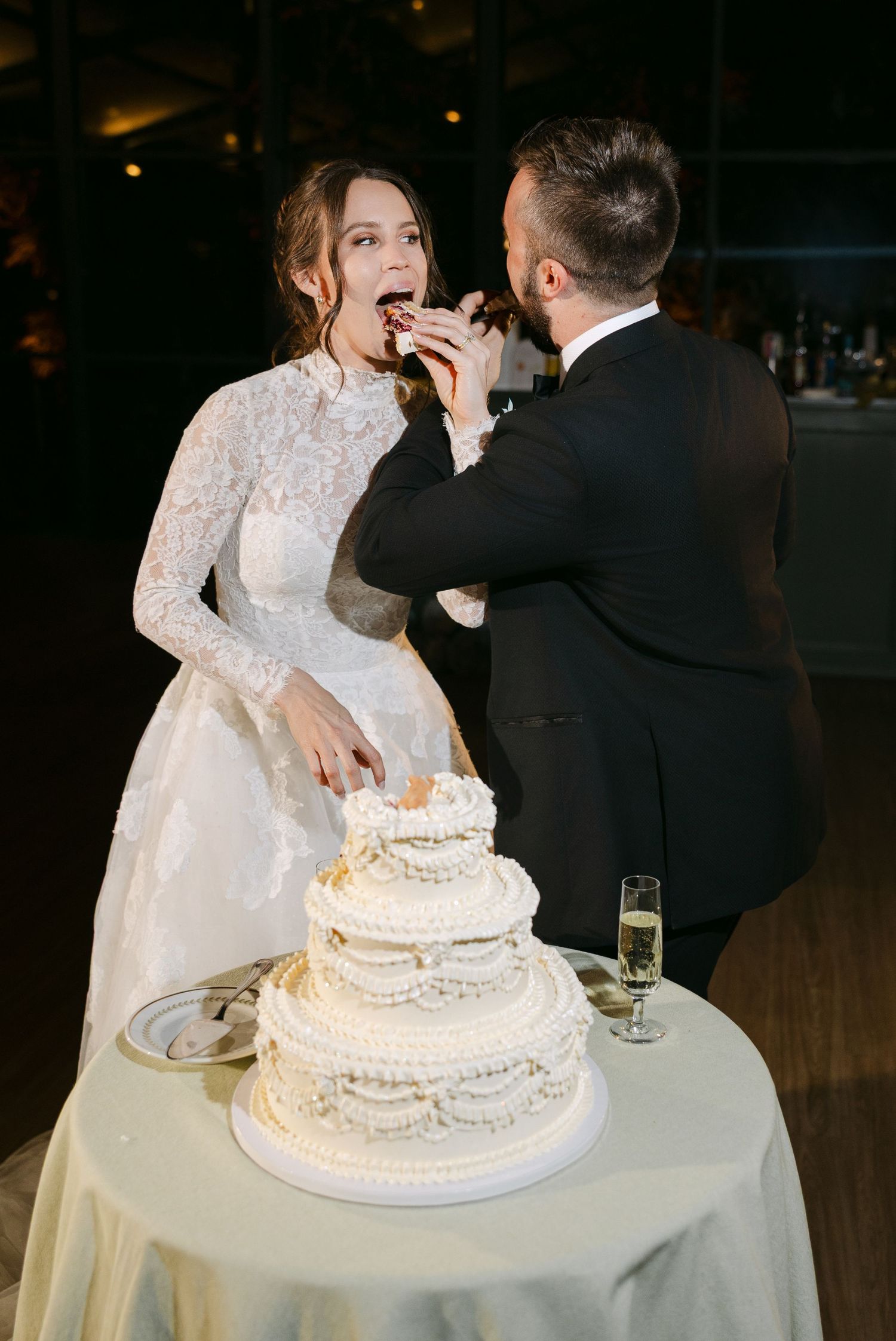 A wedding couple shares a playful moment while feeding each other cake next to their elegant white tiered wedding cake.