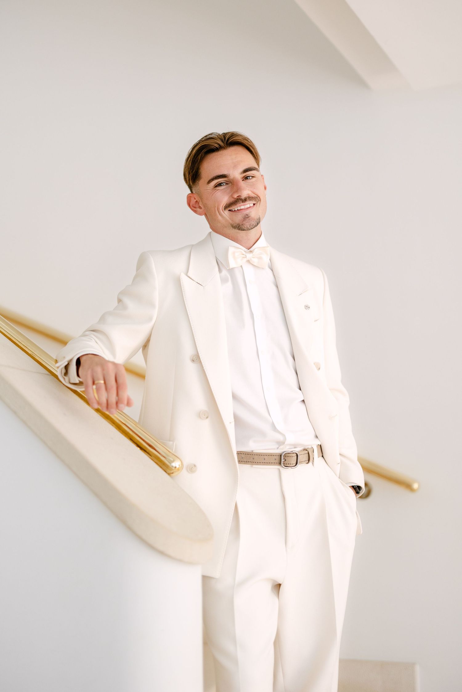 A person in a crisp white suit and bow tie poses on a staircase.